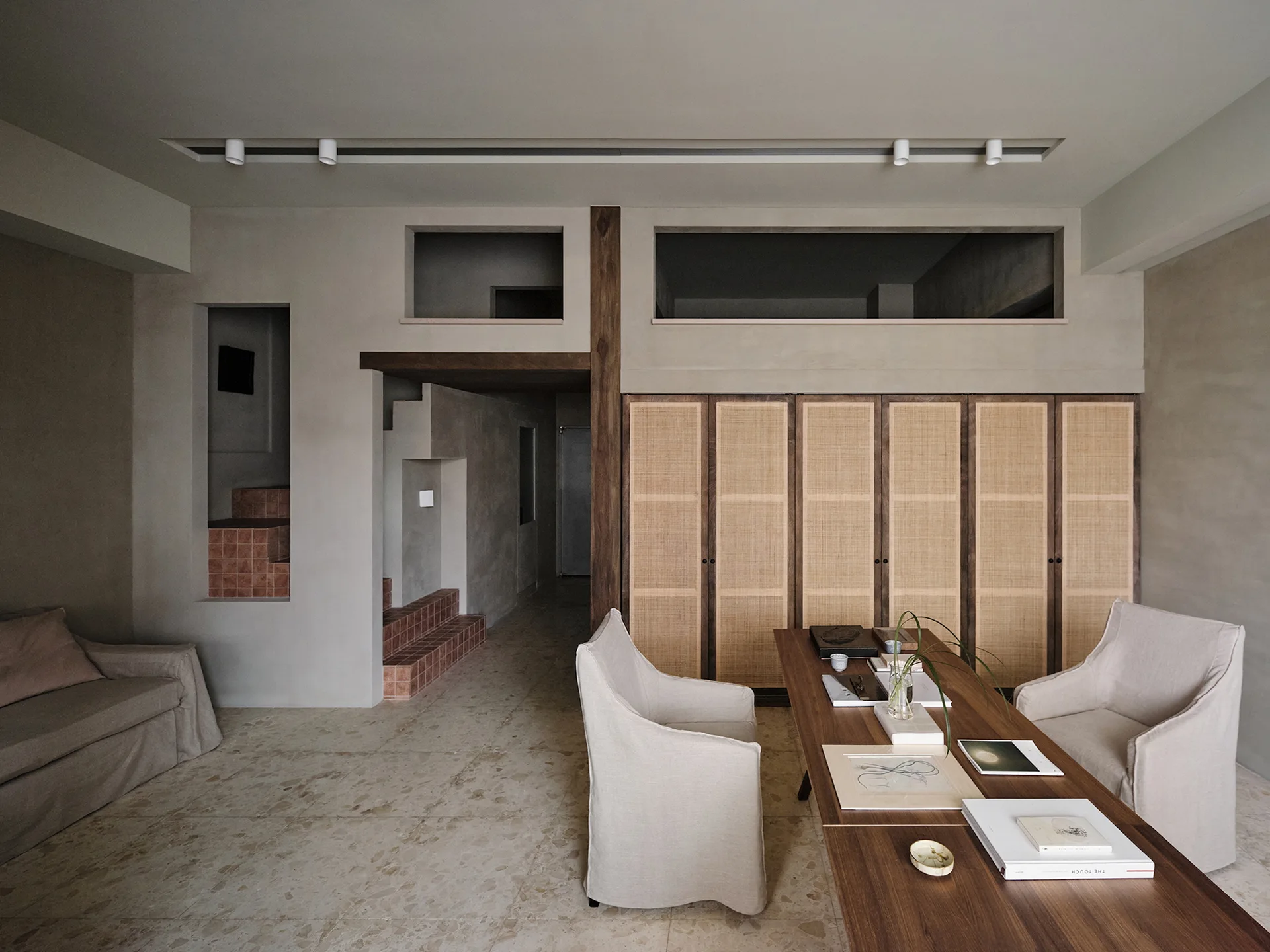 A minimalist study features a long wooden desk with two slipcovered armchairs, a large cane-front cabinet, and a glimpse of a terracotta staircase.