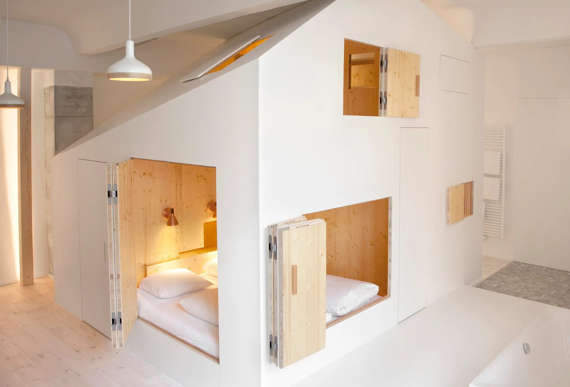 A large white built-in sleeping structure features cozy, light-wood lined alcoves with beds and folding wooden doors.