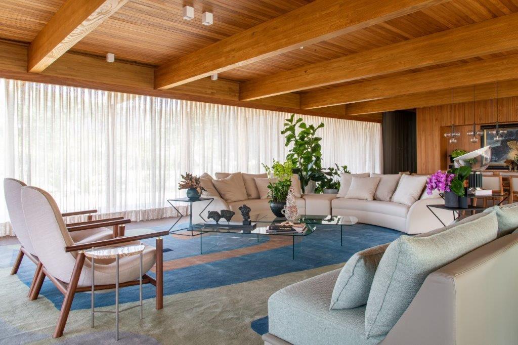 A spacious living room features a wood-beamed ceiling, cream sectional sofa, pair of wooden armchairs, and a large blue and tan patterned rug.