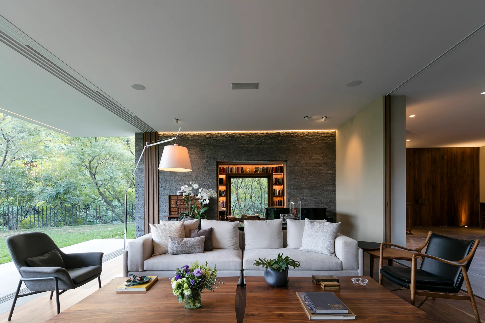 A contemporary living room with a cream sectional sofa, dual wood coffee tables, and a textured grey stone wall with a recessed bookshelf.