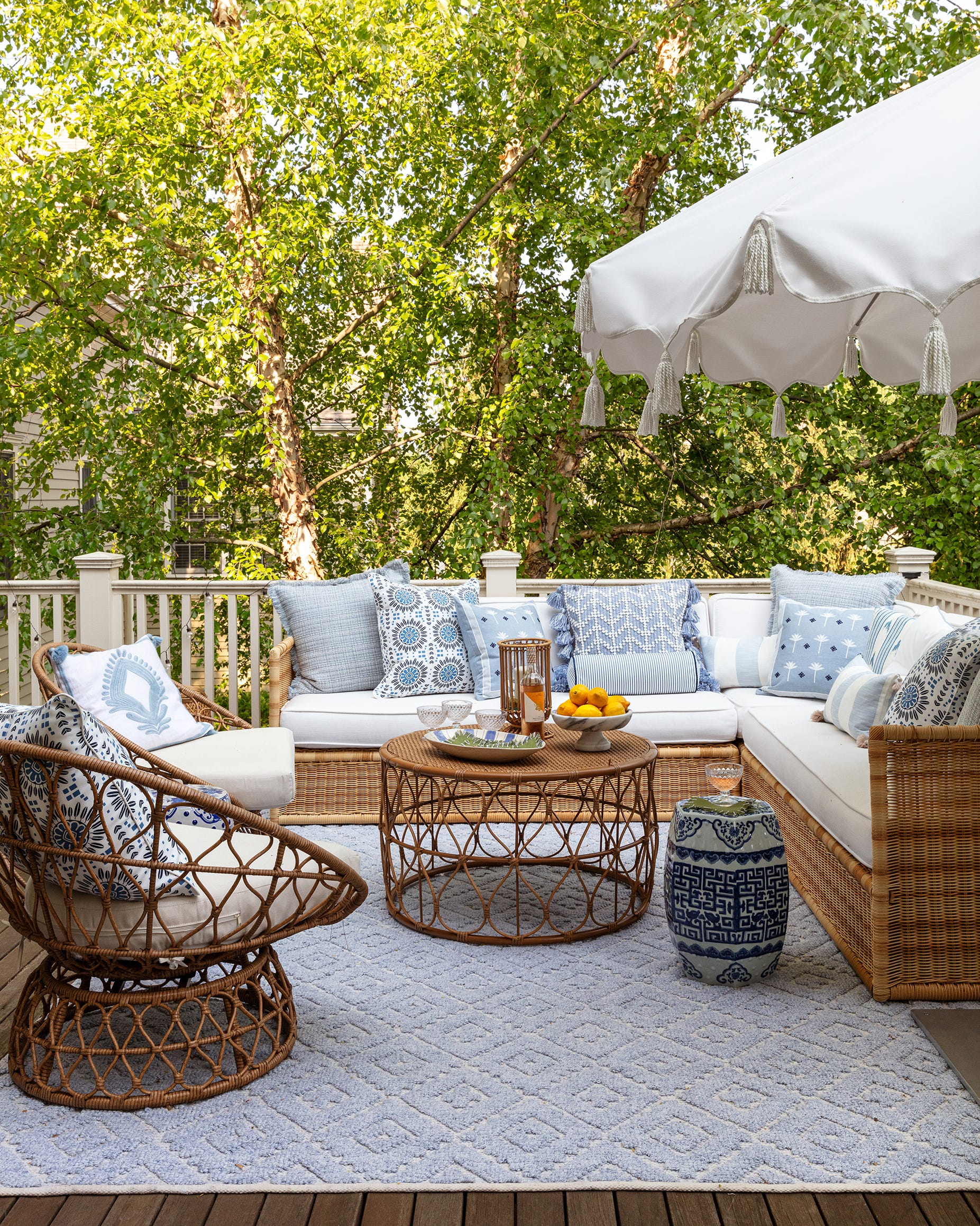 A wooden deck features a wicker sectional sofa and armchair arranged with blue patterned pillows, a round coffee table, and a tasseled white umbrella.