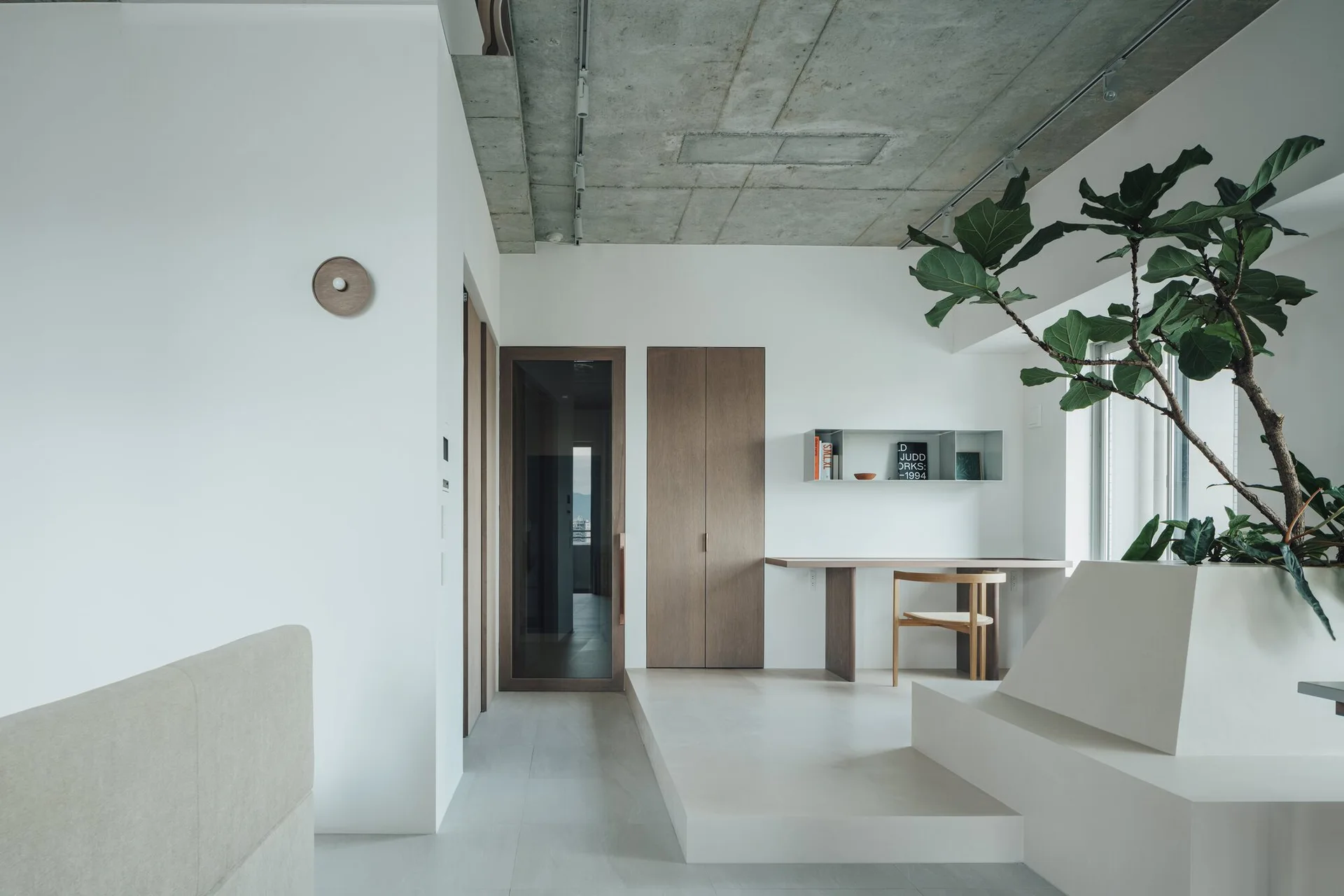 A minimalist study featuring a raised white platform, a wall-mounted wooden desk with a matching chair, and an exposed concrete ceiling.