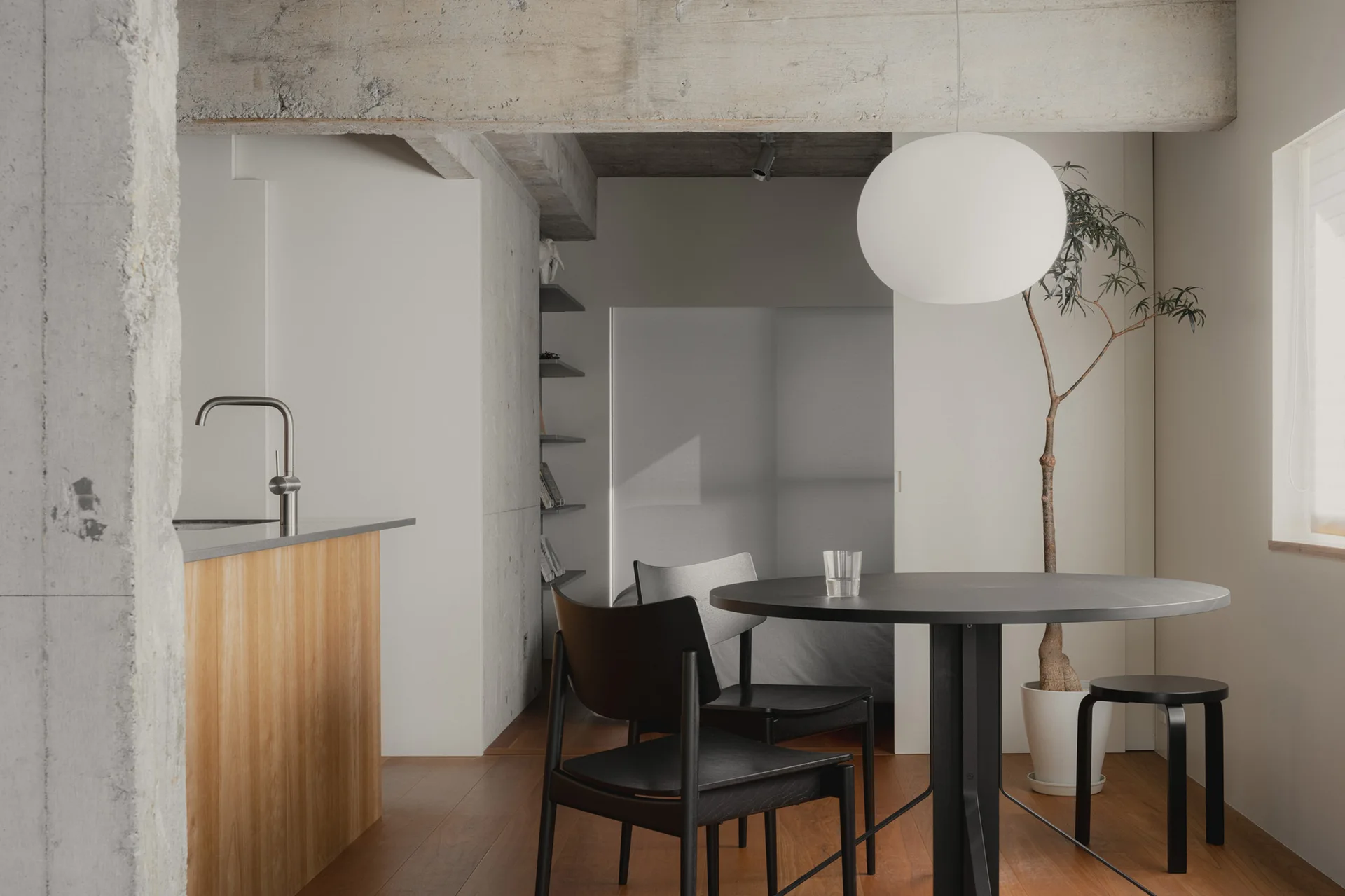 A minimalist dining space features a round black wooden table and chairs beneath an exposed concrete ceiling beam and a large white globe pendant light.