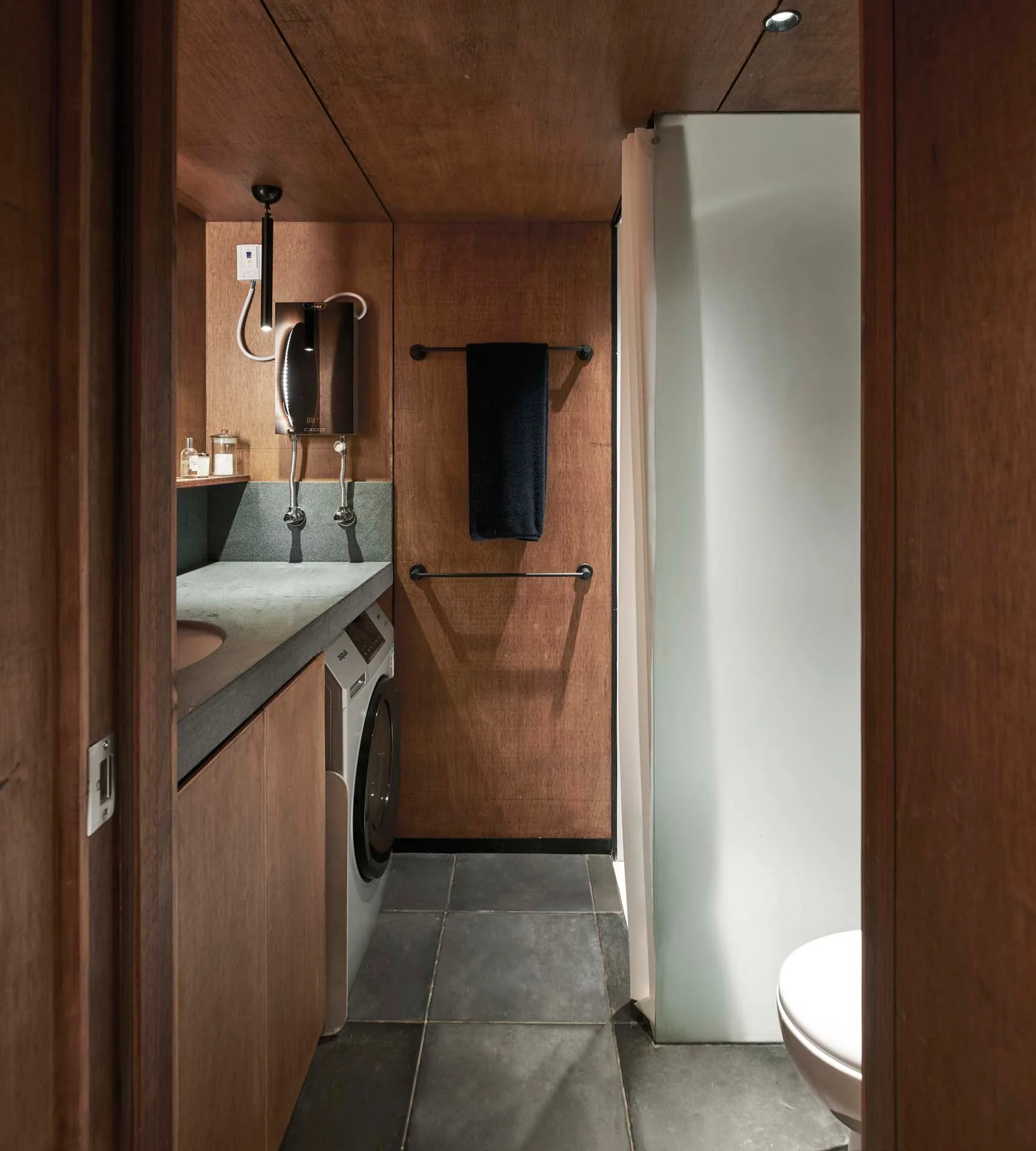A compact modern bathroom with dark wood-paneled walls, gray stone floor tiles, and a washing machine integrated into the wood vanity.