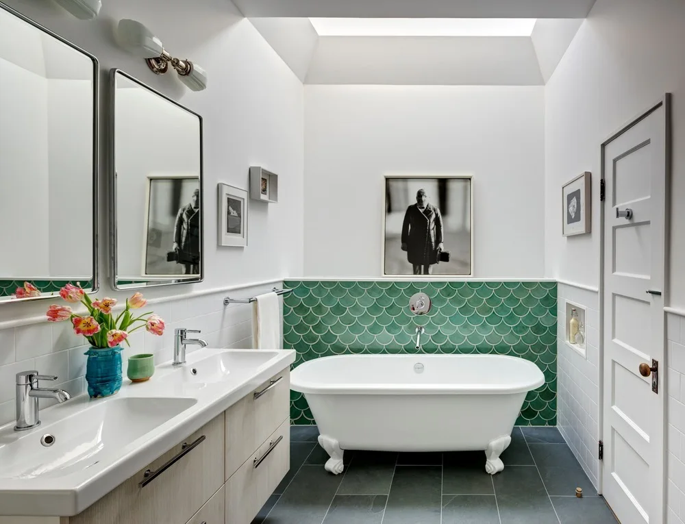 A modern bathroom features a white clawfoot bathtub set against a green scalloped tile wall, beside a light wood double vanity and under a skylight.