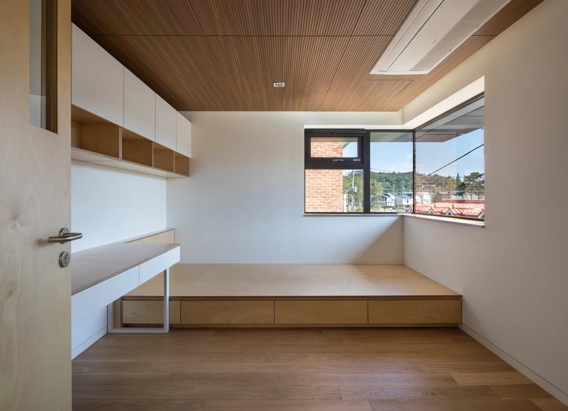 A minimalist bedroom featuring a built-in plywood platform bed with drawers, an integrated desk, white upper cabinets, and a wood slat ceiling.