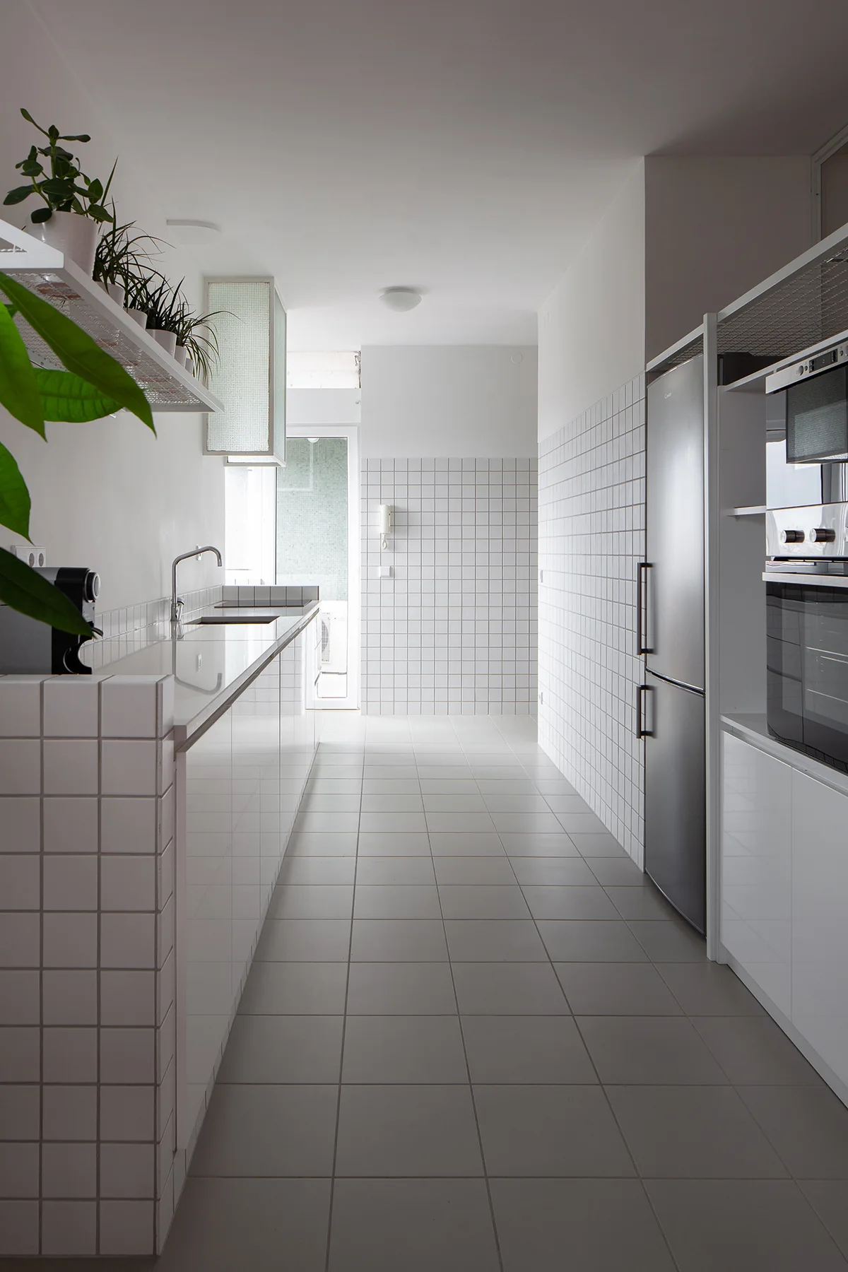 A narrow galley kitchen featuring white square-tiled walls and counters, a gray tiled floor, a stainless steel refrigerator, and open shelving.