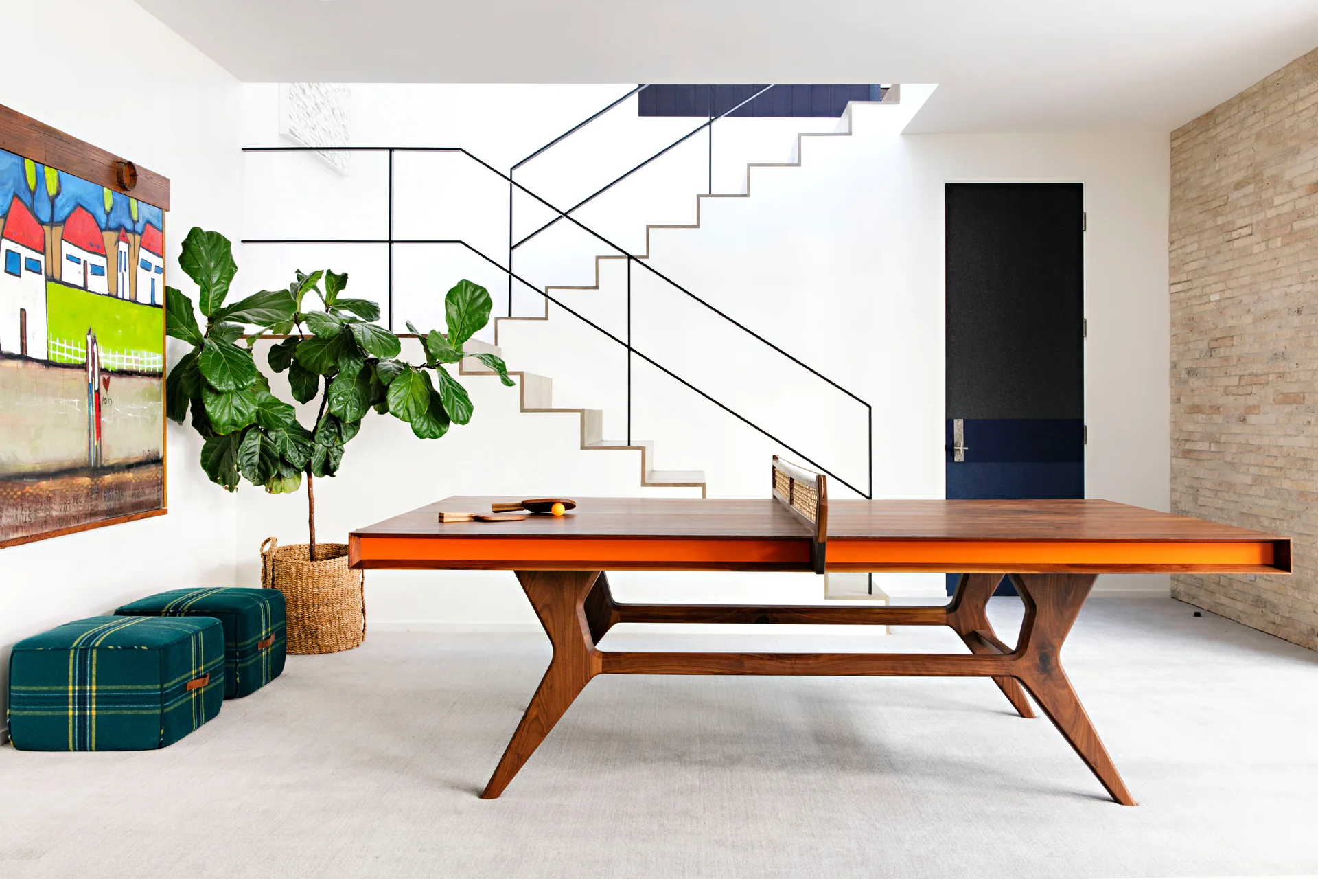 A modern recreation room features a wooden ping pong table with an orange edge, green plaid ottomans, and an open wood staircase.