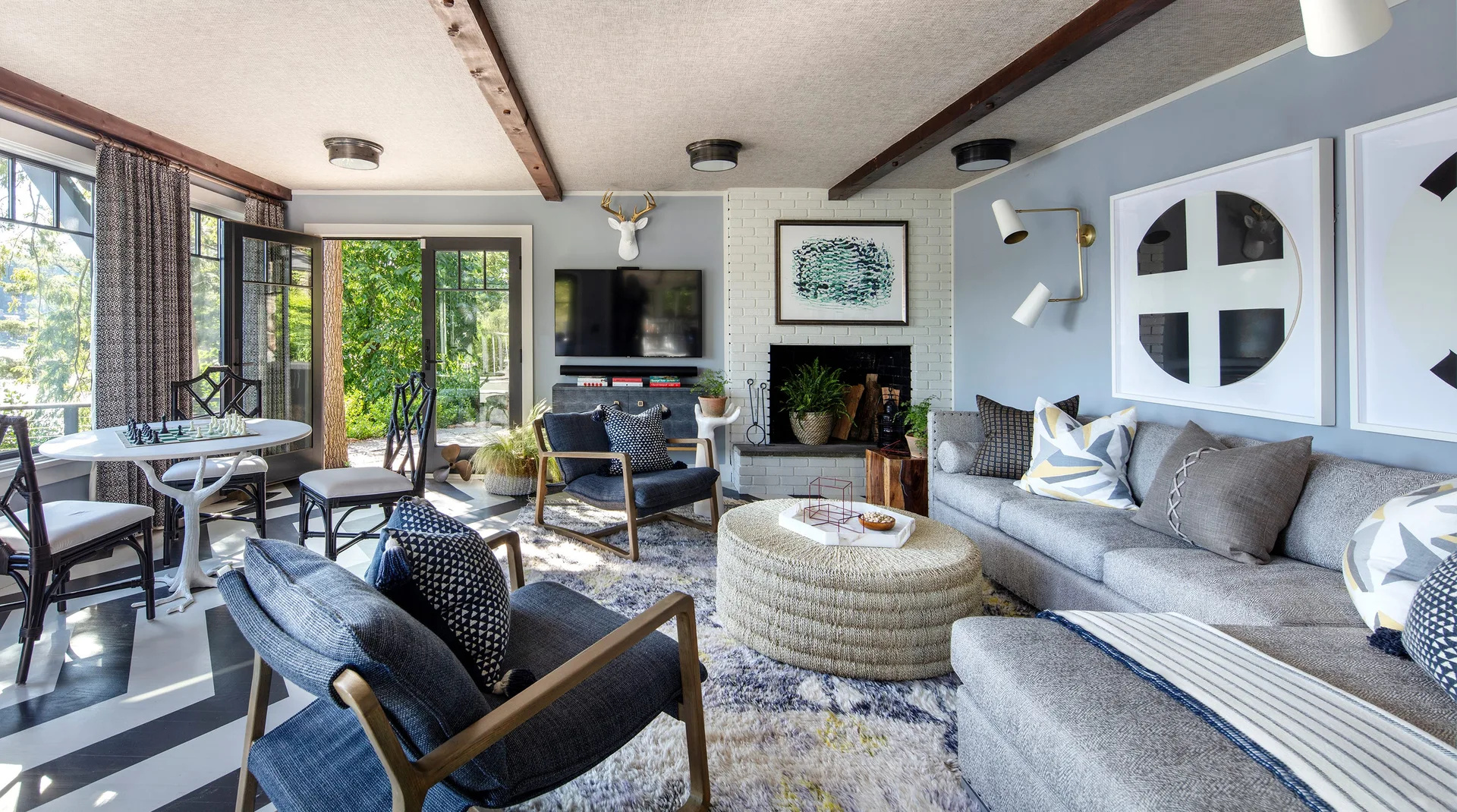 A transitional living room features a grey sectional, blue armchairs, a woven round ottoman, ceiling beams, and a black-and-white chevron floor.