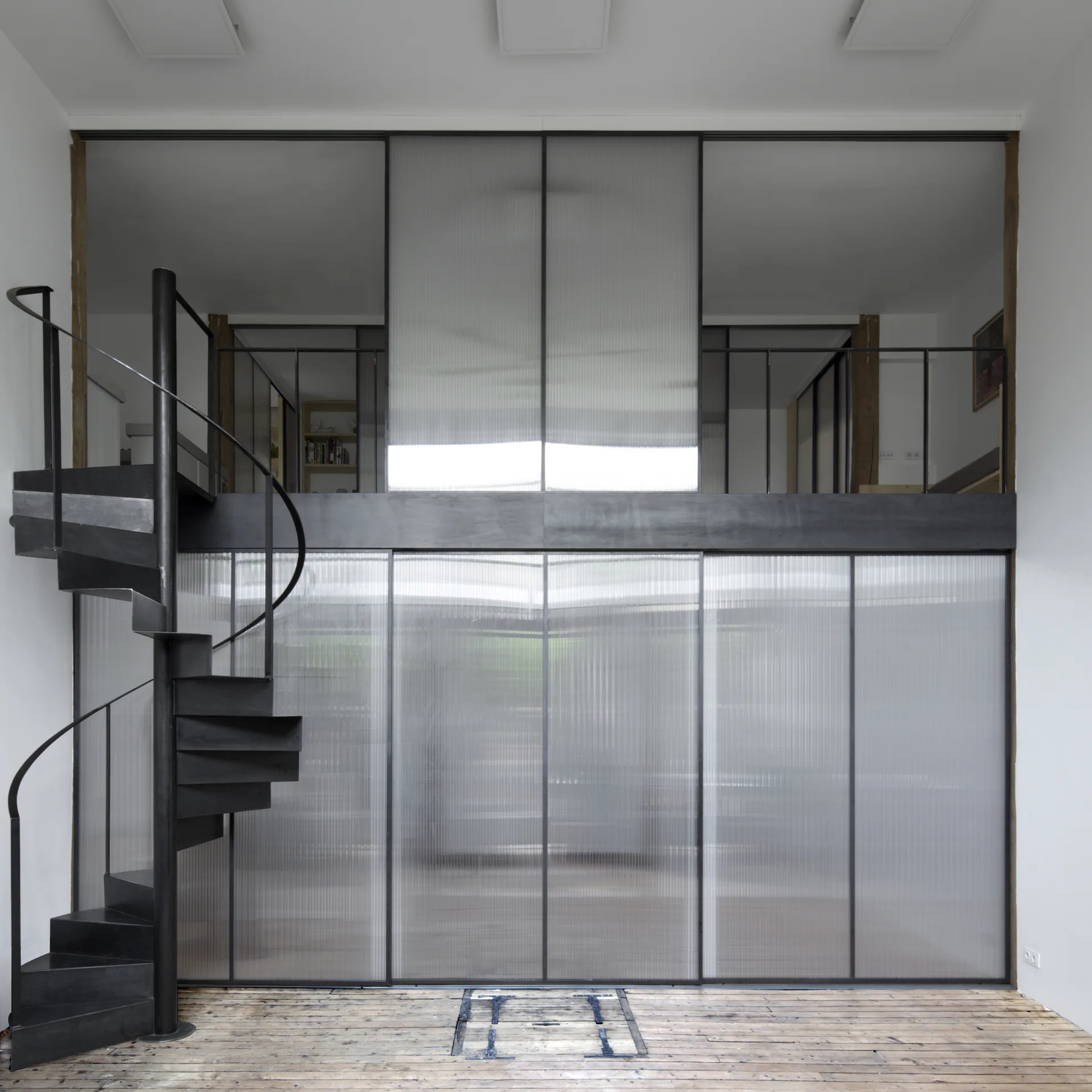 A black metal spiral staircase stands next to a two-story translucent ribbed partition wall inside a minimalist loft with wood floors.
