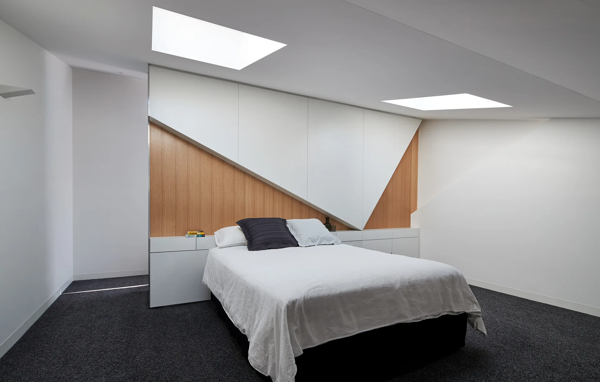 A minimalist bedroom featuring a bed centered against an angular white and wood built-in cabinet system beneath two ceiling skylights.