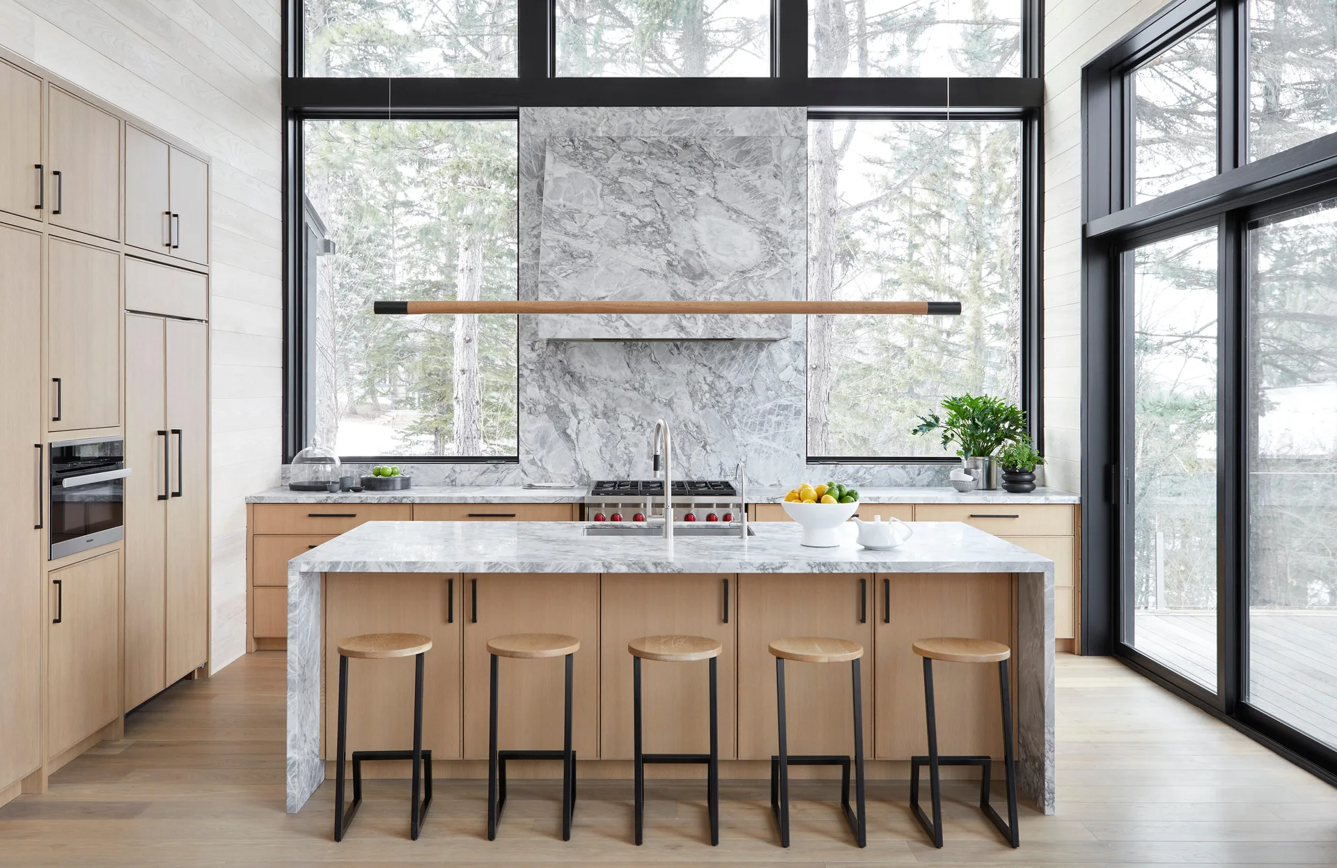 A modern kitchen featuring light wood cabinetry, a marble waterfall island with bar stools, and large black-framed windows overlooking pine trees.