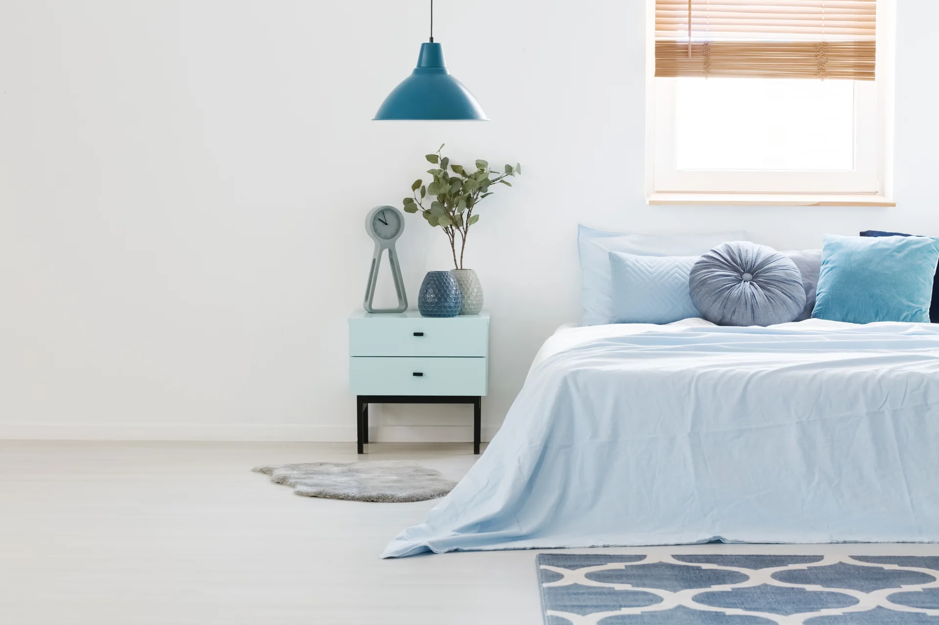 A minimalist bedroom featuring light blue bedding, a matching two-drawer nightstand, and a dark blue pendant light hanging against a white wall.