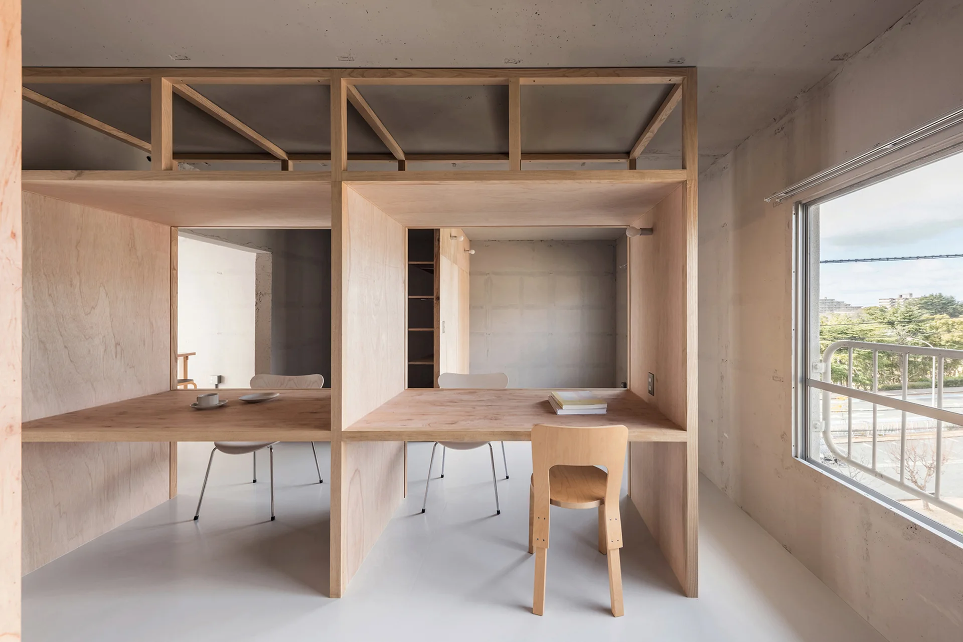 A minimalist home office featuring built-in plywood desk cubicles with chairs next to a large window and raw concrete walls.