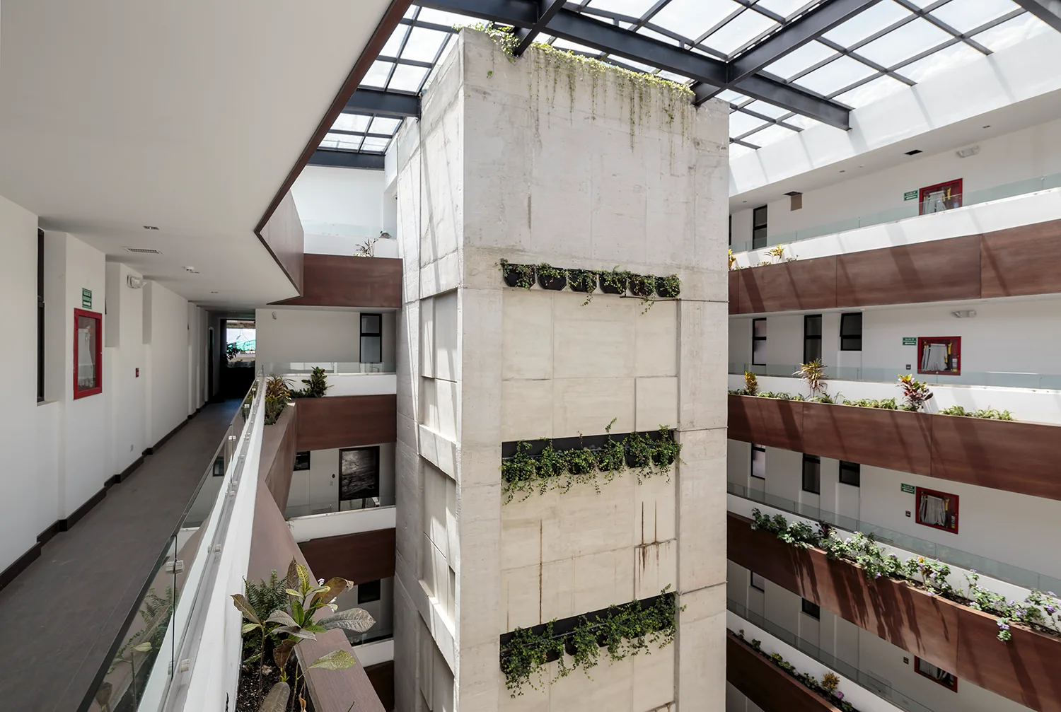A multi-story indoor atrium features a central concrete elevator tower with horizontal planters under a large grid skylight roof.
