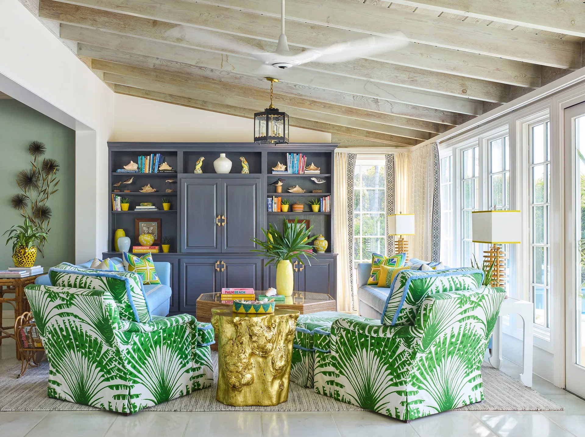 A bright living room features light blue sofas, vibrant palm-print armchairs, and a large grey built-in bookcase under a vaulted wood ceiling.