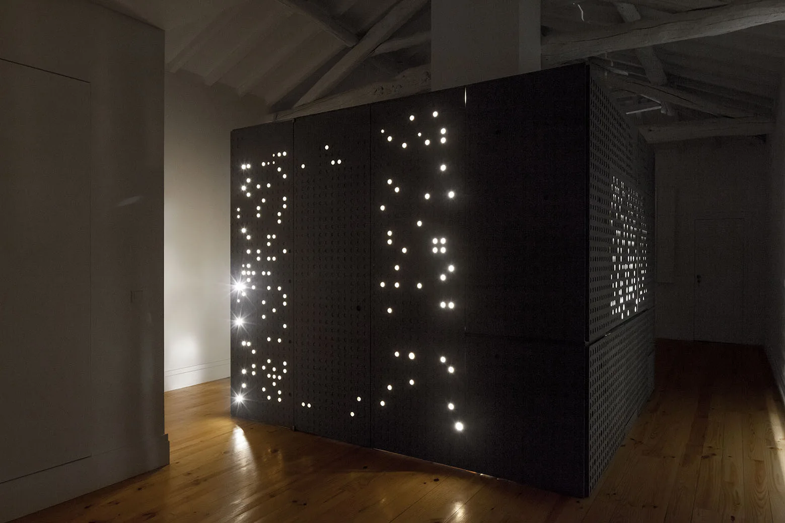 A large matte black perforated cabinet with integrated lighting stands in a room with warm hardwood floors and exposed white ceiling beams.