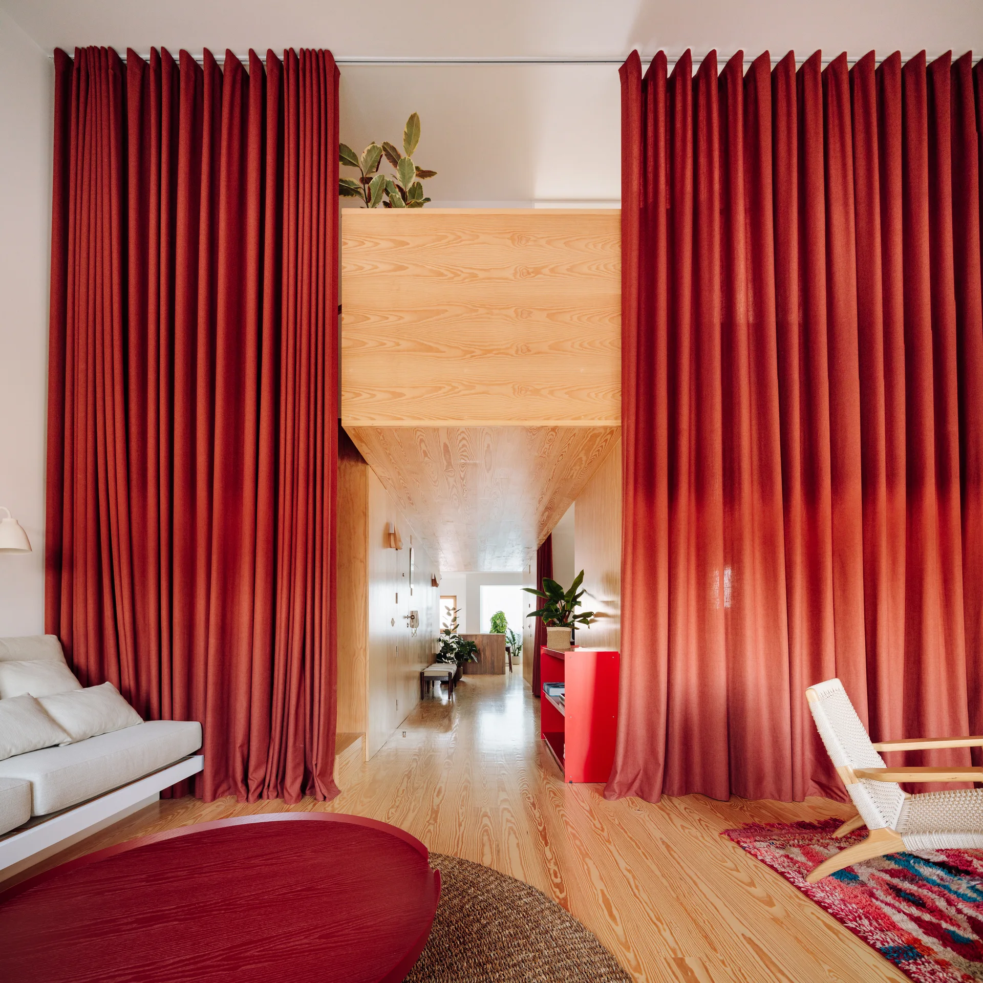 Floor-to-ceiling red curtains frame a light wood-clad hallway structure in a modern living room with a white sofa and woven chair.