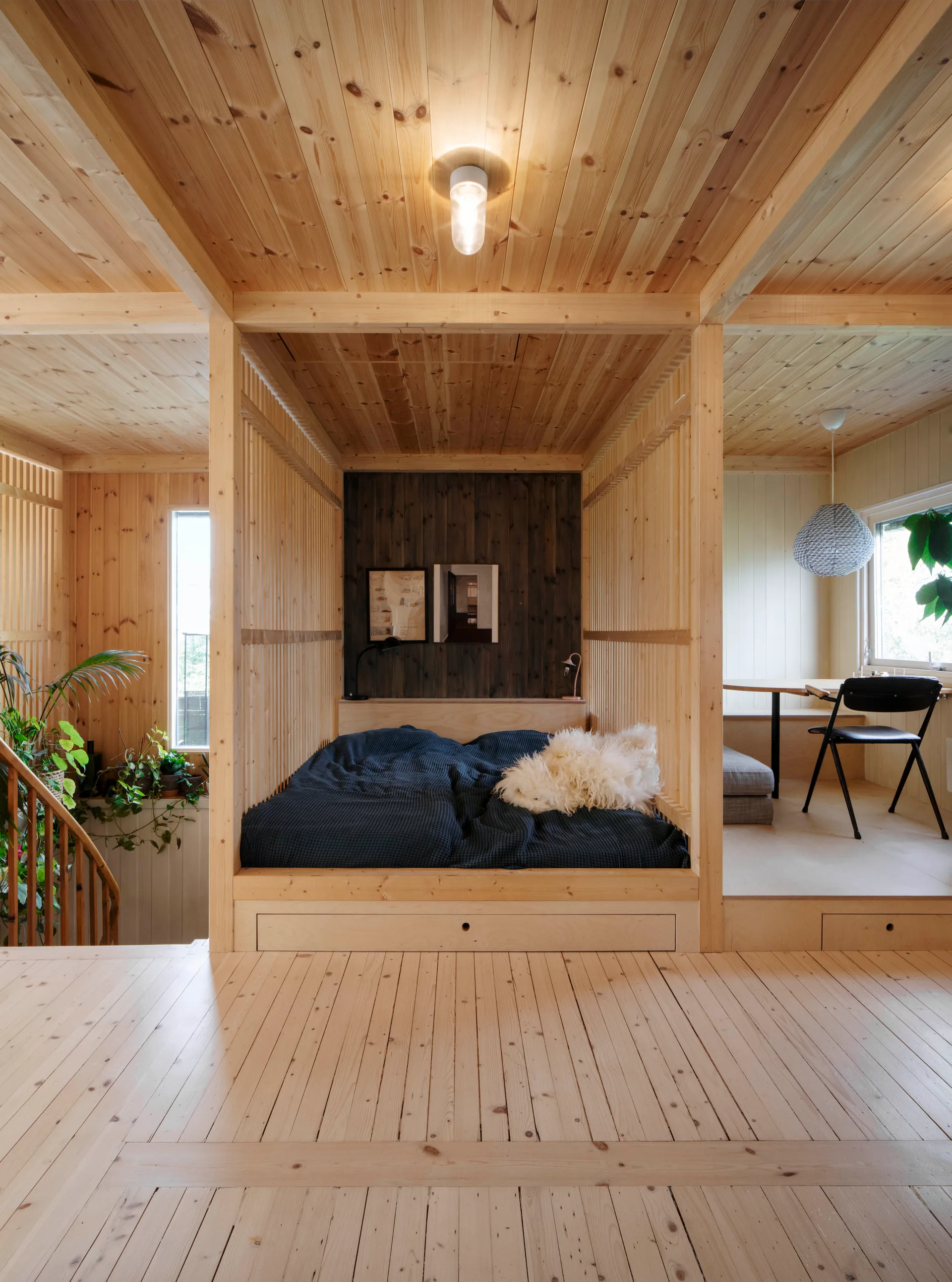 A built-in platform bed is enclosed by a vertical slatted pine frame, next to a raised dining area under a wood plank ceiling.