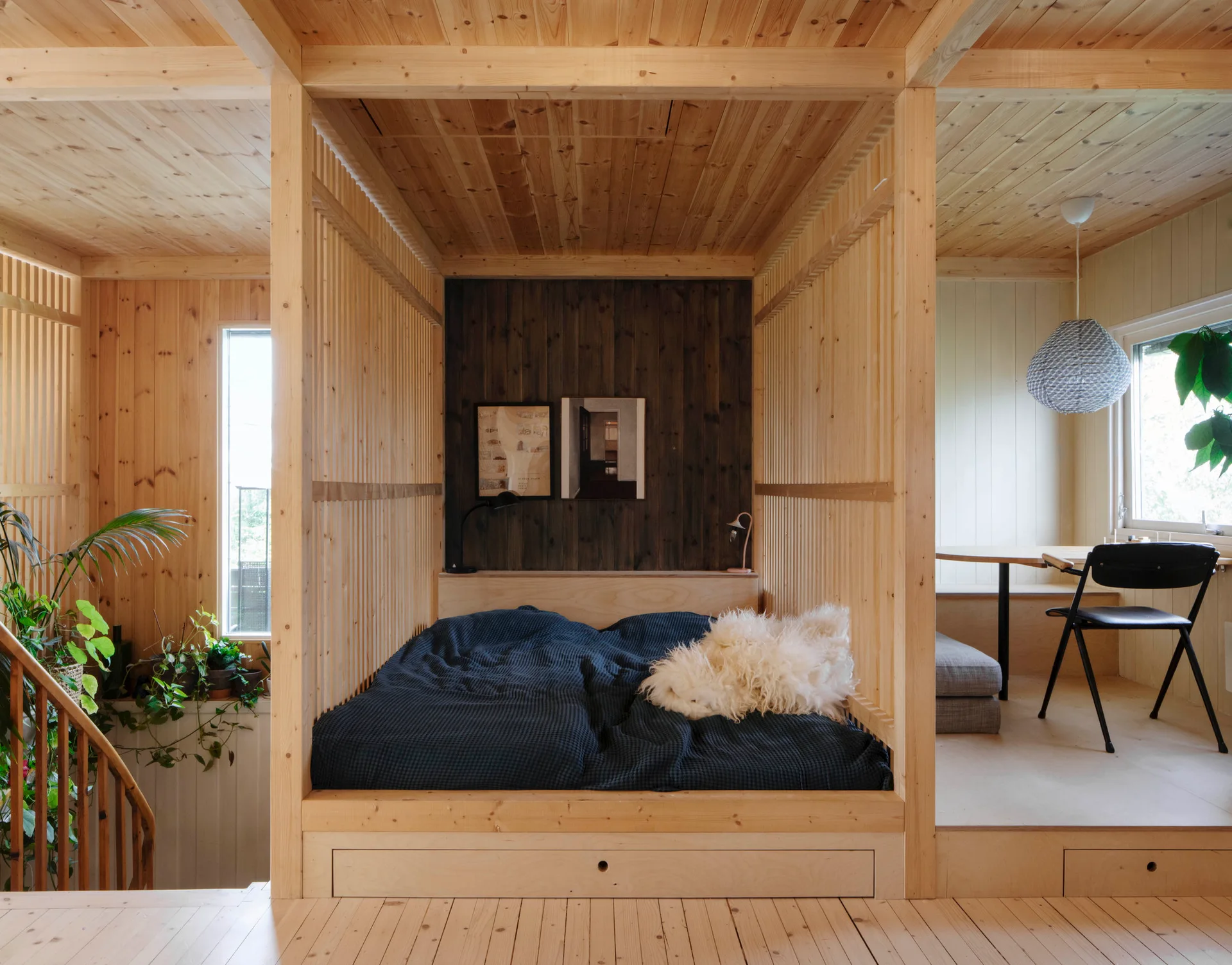 A built-in platform bed with dark blue bedding is enclosed by vertical wood slats in a light wood cabin bedroom with a split-level floor.