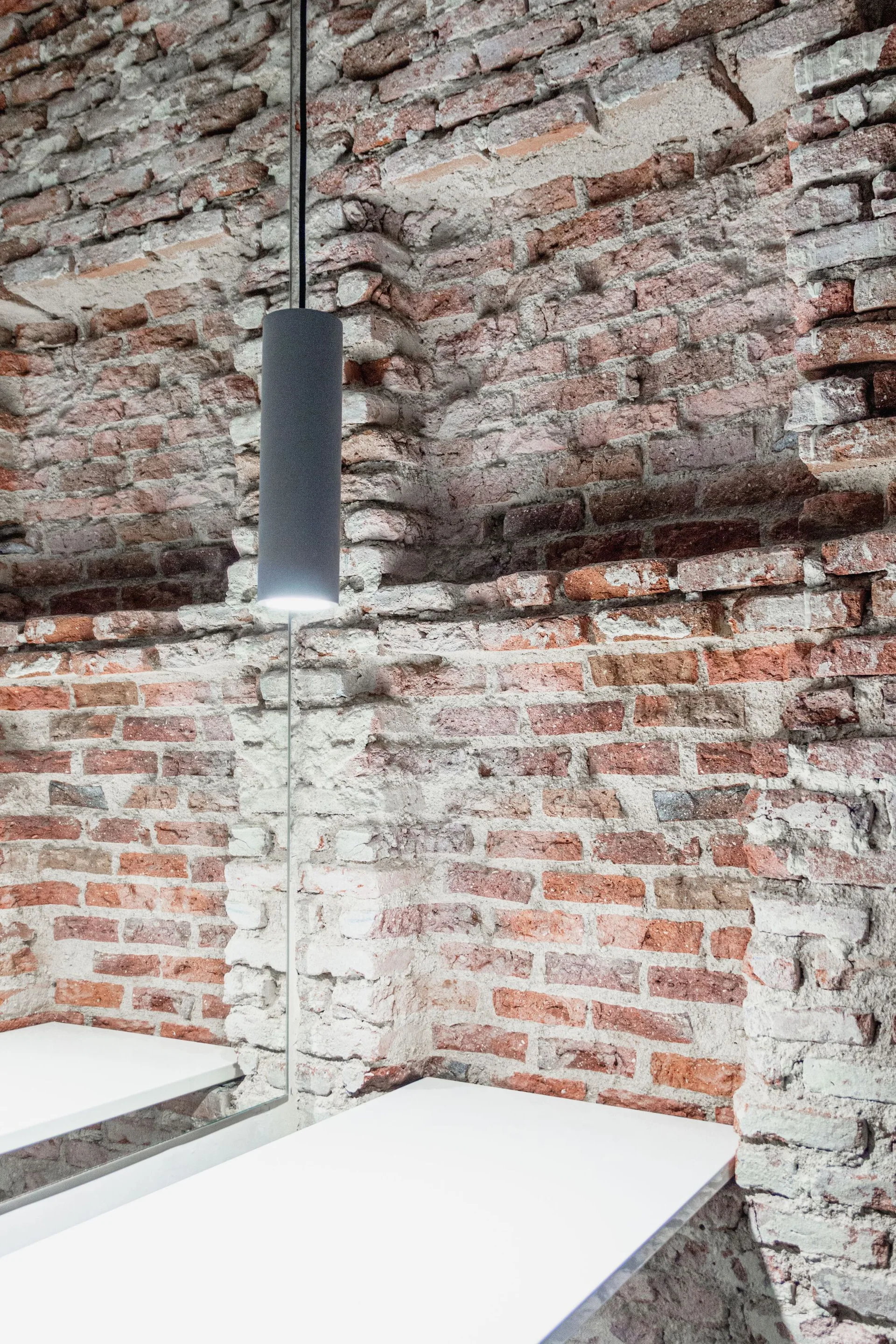 A cylindrical dark pendant light hangs in front of a textured red brick wall above a clean white surface reflected in a mirror.