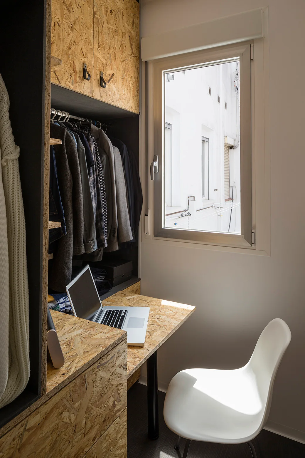 A compact workspace features an integrated desk and wardrobe made of oriented strand board next to a white plastic chair and a window.