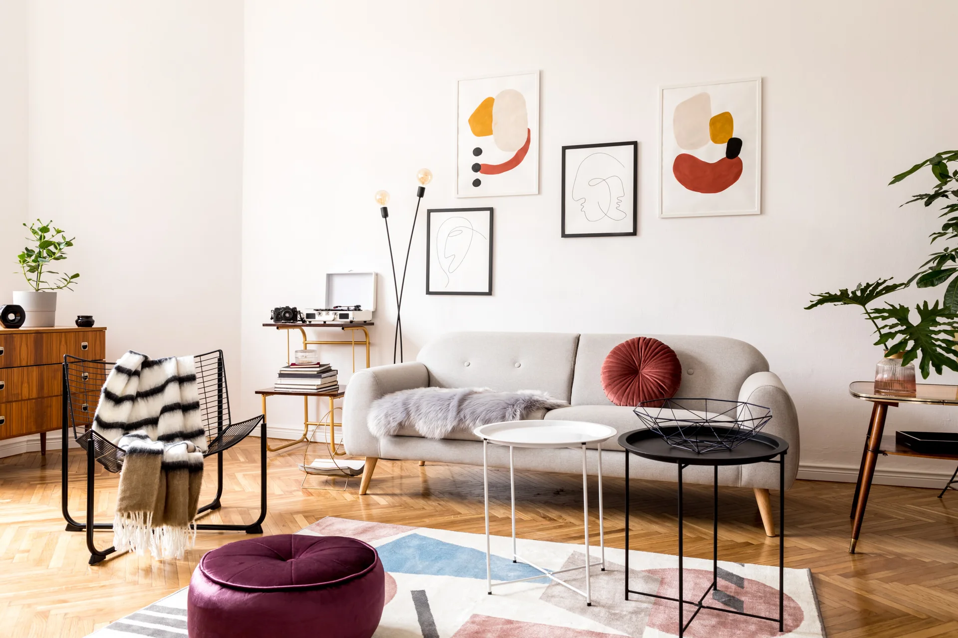 A light gray sofa sits on a herringbone wood floor in a living room decorated with a geometric rug, colorful abstract art, and a black wire chair.