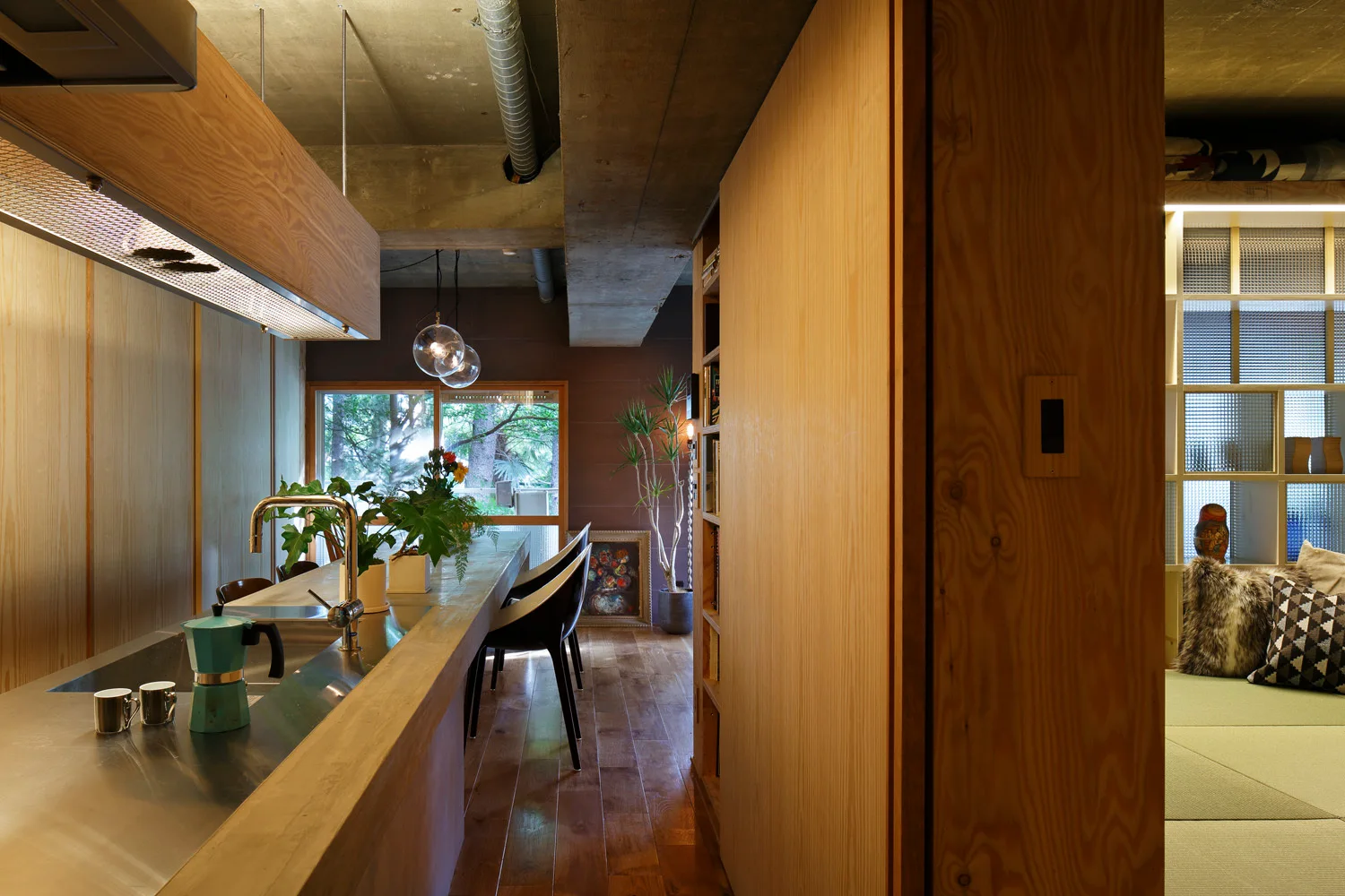 A modern kitchen featuring a long wood and stainless steel island, wood-paneled walls, an exposed concrete ceiling, and black dining chairs.