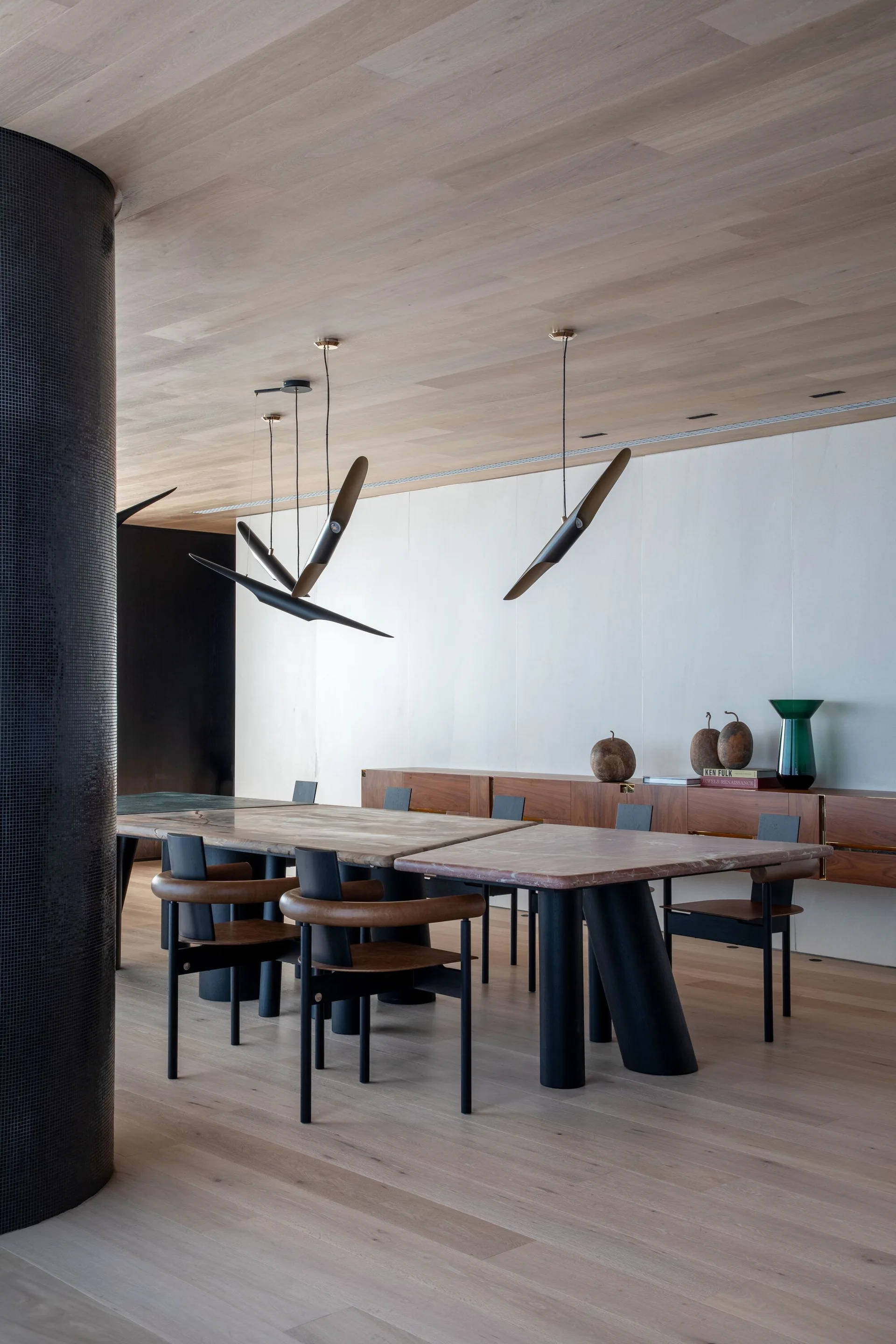 A modern dining room features a large stone-topped table, curved dark armchairs, sculptural pendant lights, and a matching wood floor and ceiling.