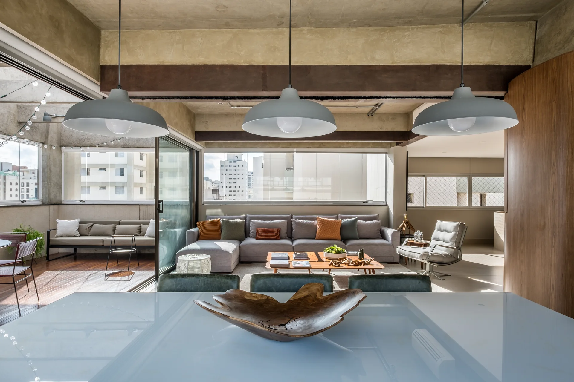 A modern living room with a grey sectional sofa, low wood coffee tables, three large pendant lights, and a balcony visible through sliding doors.