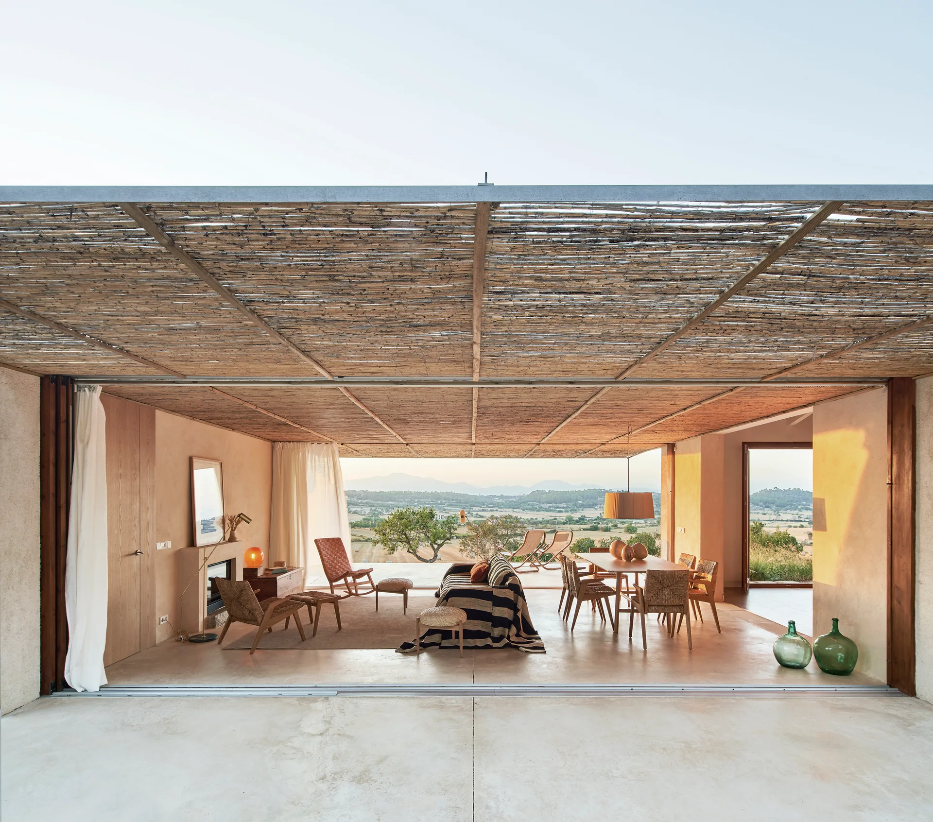 An open-plan living and dining room features a woven reed ceiling, a striped sofa, a wooden dining table, and wide openings leading to an outdoor terrace.