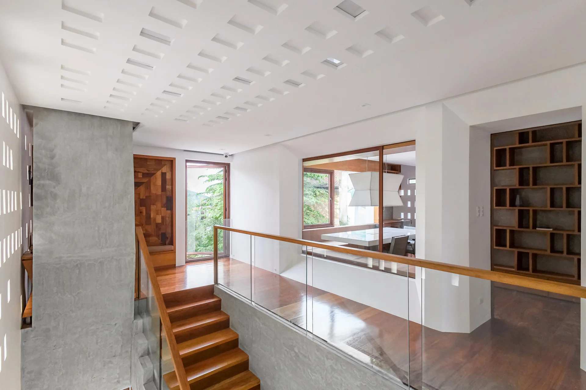 A modern home landing features polished wood floors, a staircase with a glass railing, concrete walls, and a recessed grid patterned white ceiling.