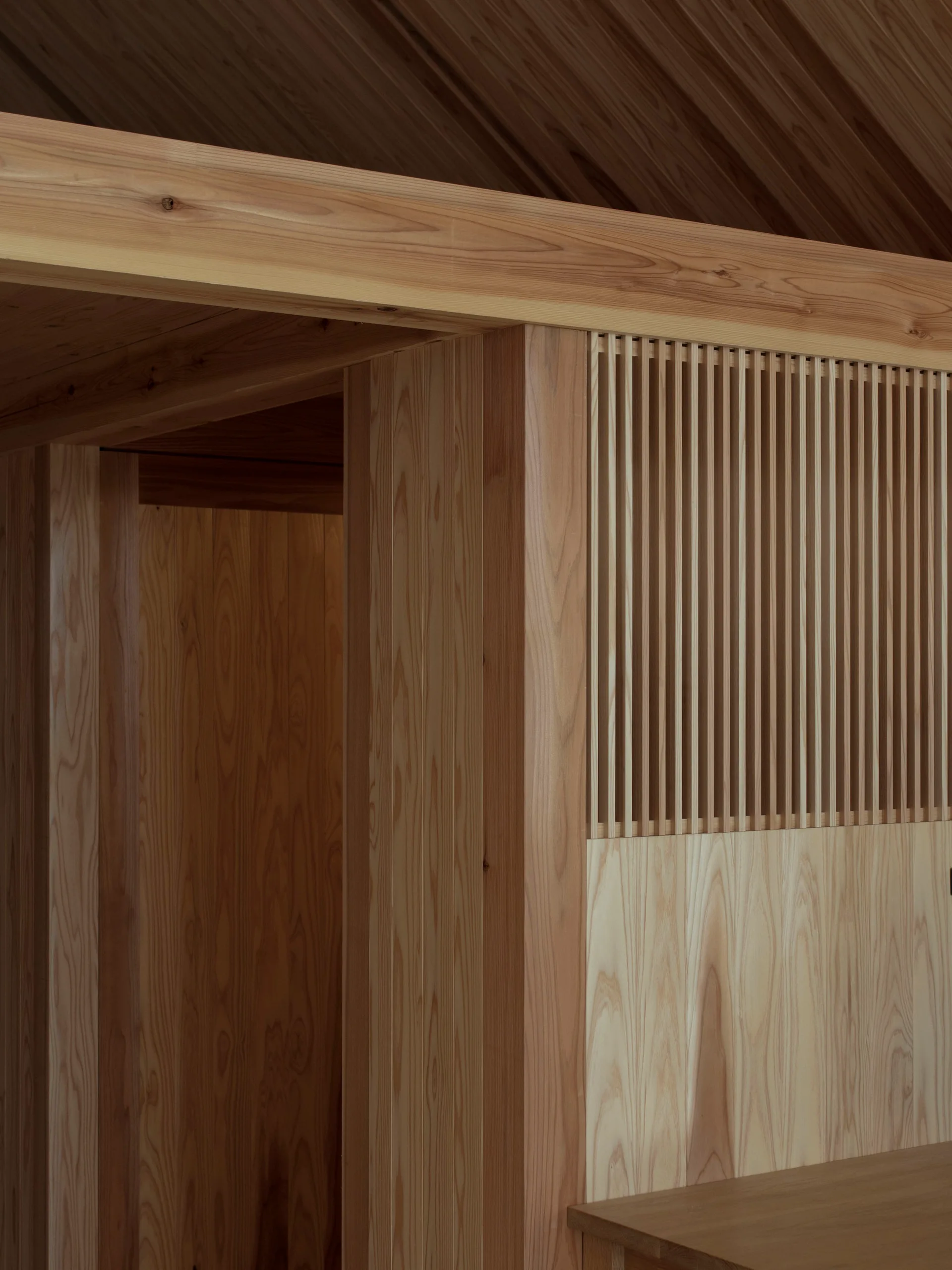 A close-up detail of a minimalist room showing natural wood paneled walls, a large timber beam, and a vertical slatted screen partition.