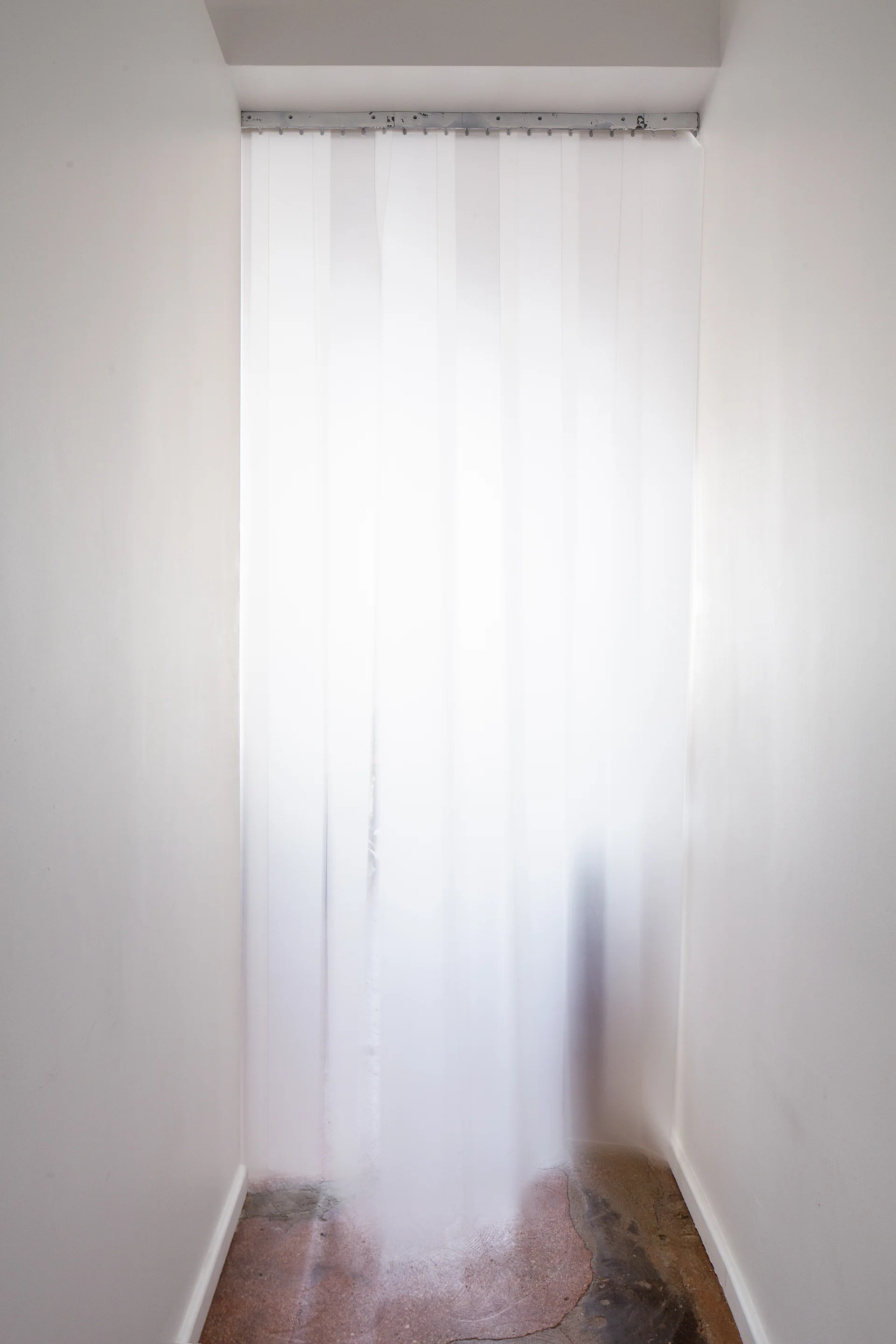 A translucent white plastic strip curtain hangs from a metal track in a narrow white-walled doorway with a concrete floor.
