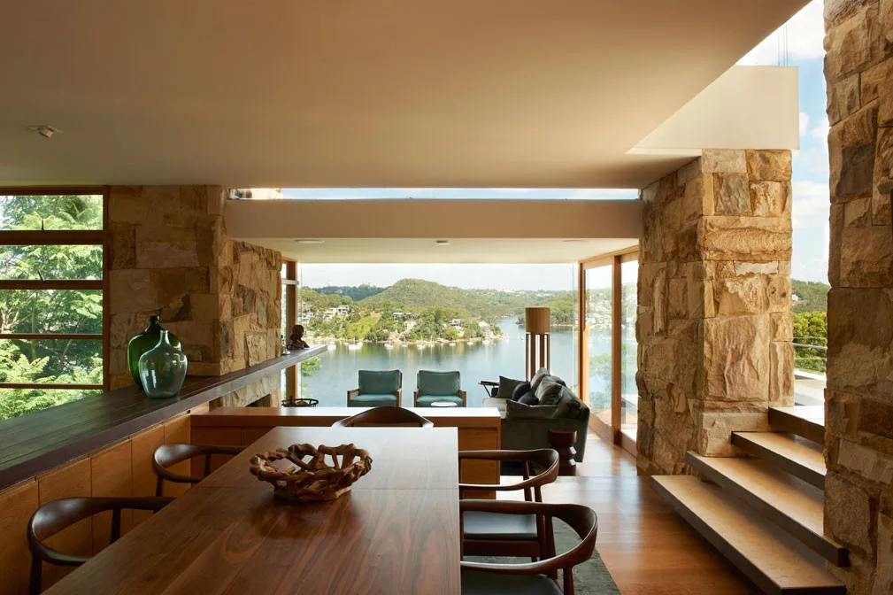 An open-plan dining and living room features exposed stone walls, a wooden dining table, and large windows showcasing a waterfront view.