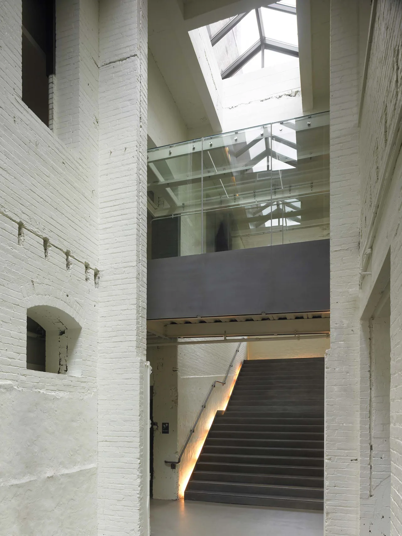 A dark modern staircase sits between tall painted white brick walls under an overhead glass bridge and skylight.