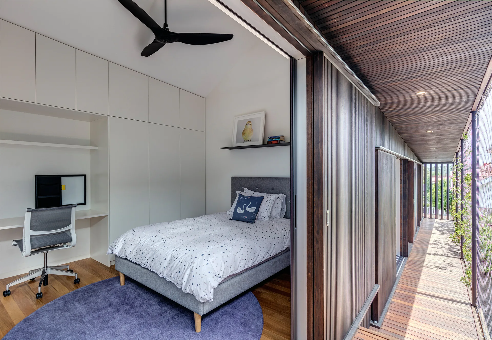 A compact bedroom features a grey single bed, built-in white desk with chair, and a slatted sliding wood door that opens to an outdoor wooden walkway.