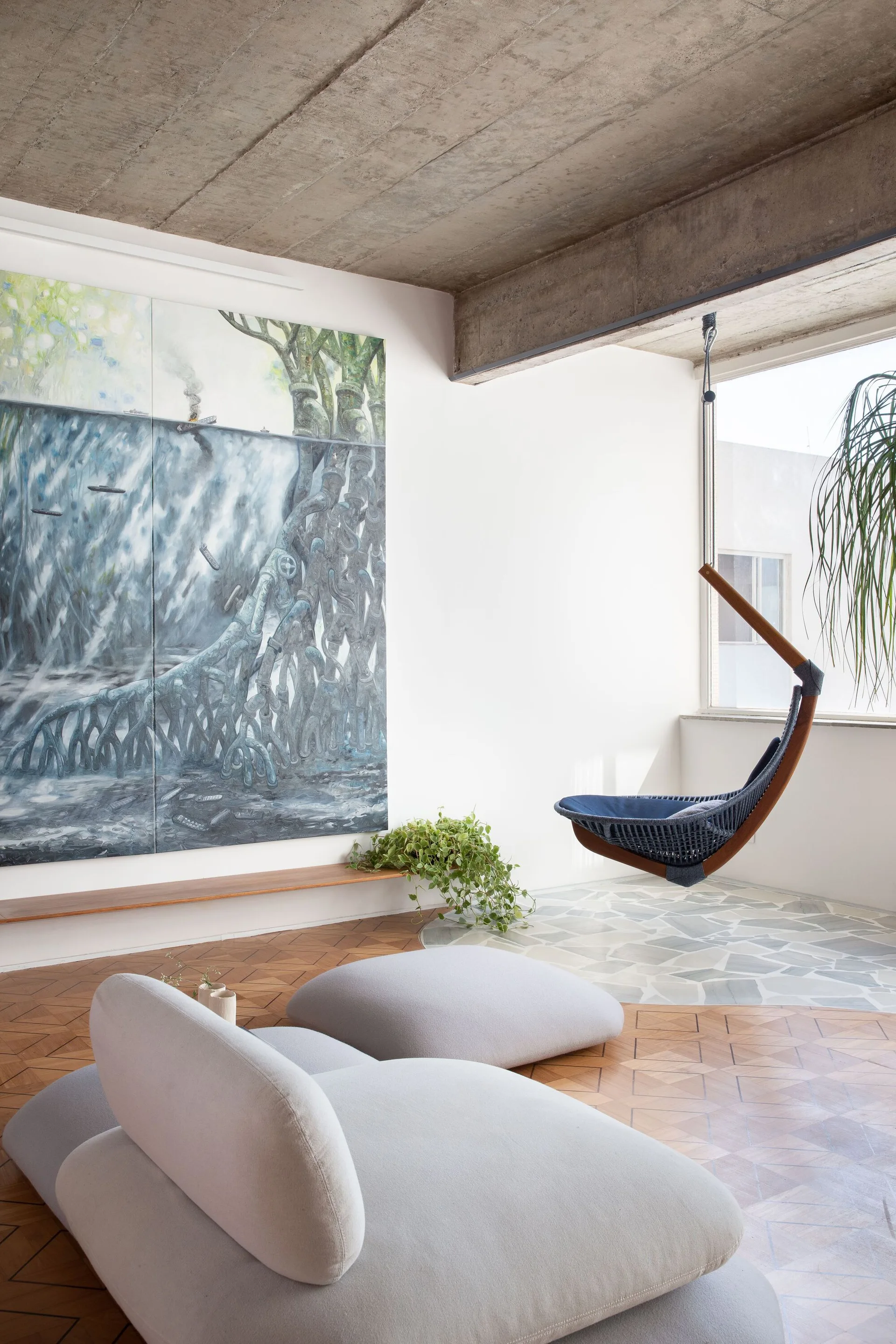 A modern living space features a low-profile grey sofa, a hanging chair near a window, an exposed concrete ceiling, and a large wall painting.