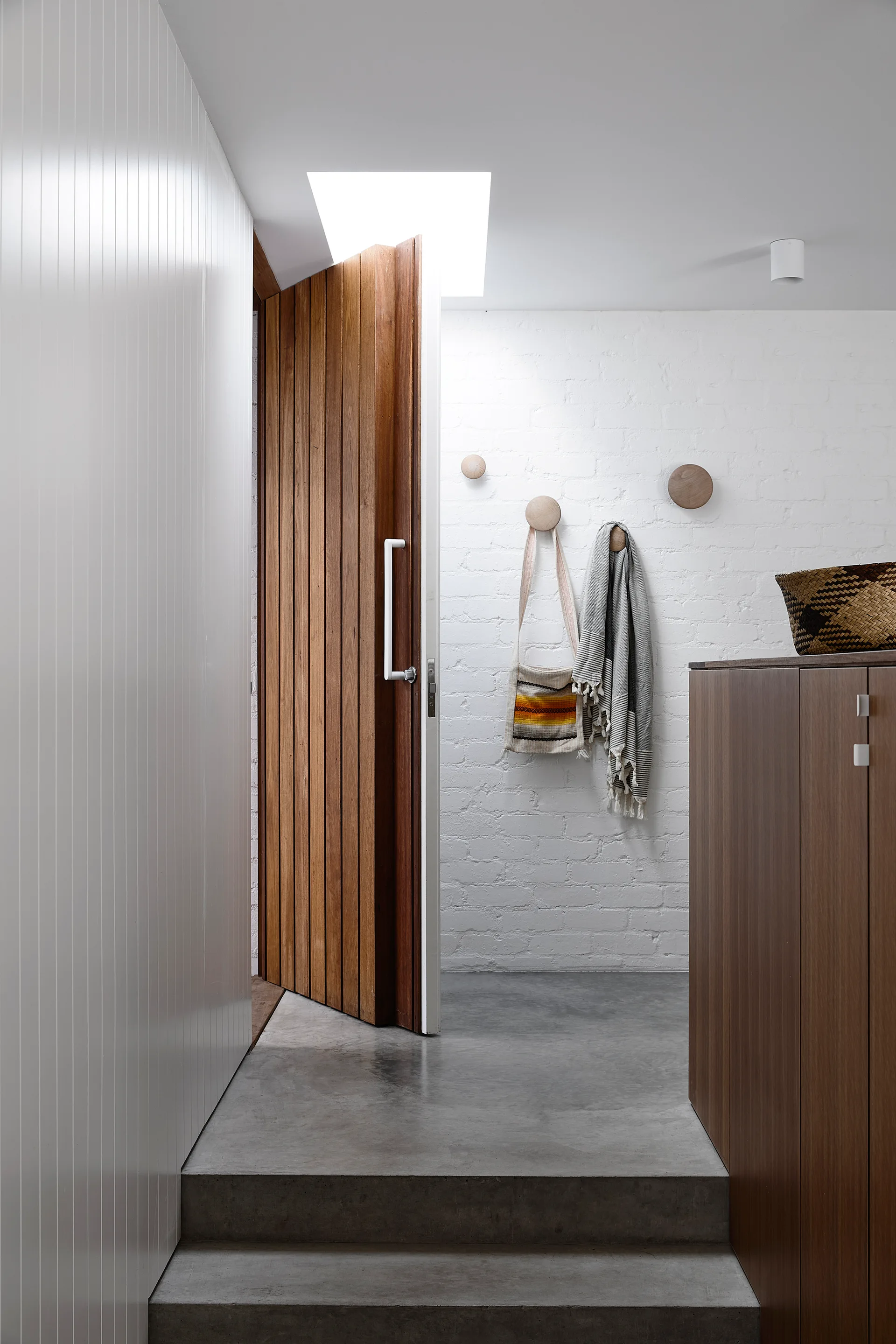 A modern entryway features a slatted wood door under a skylight, with concrete steps, a painted brick wall, and round wooden wall hooks.