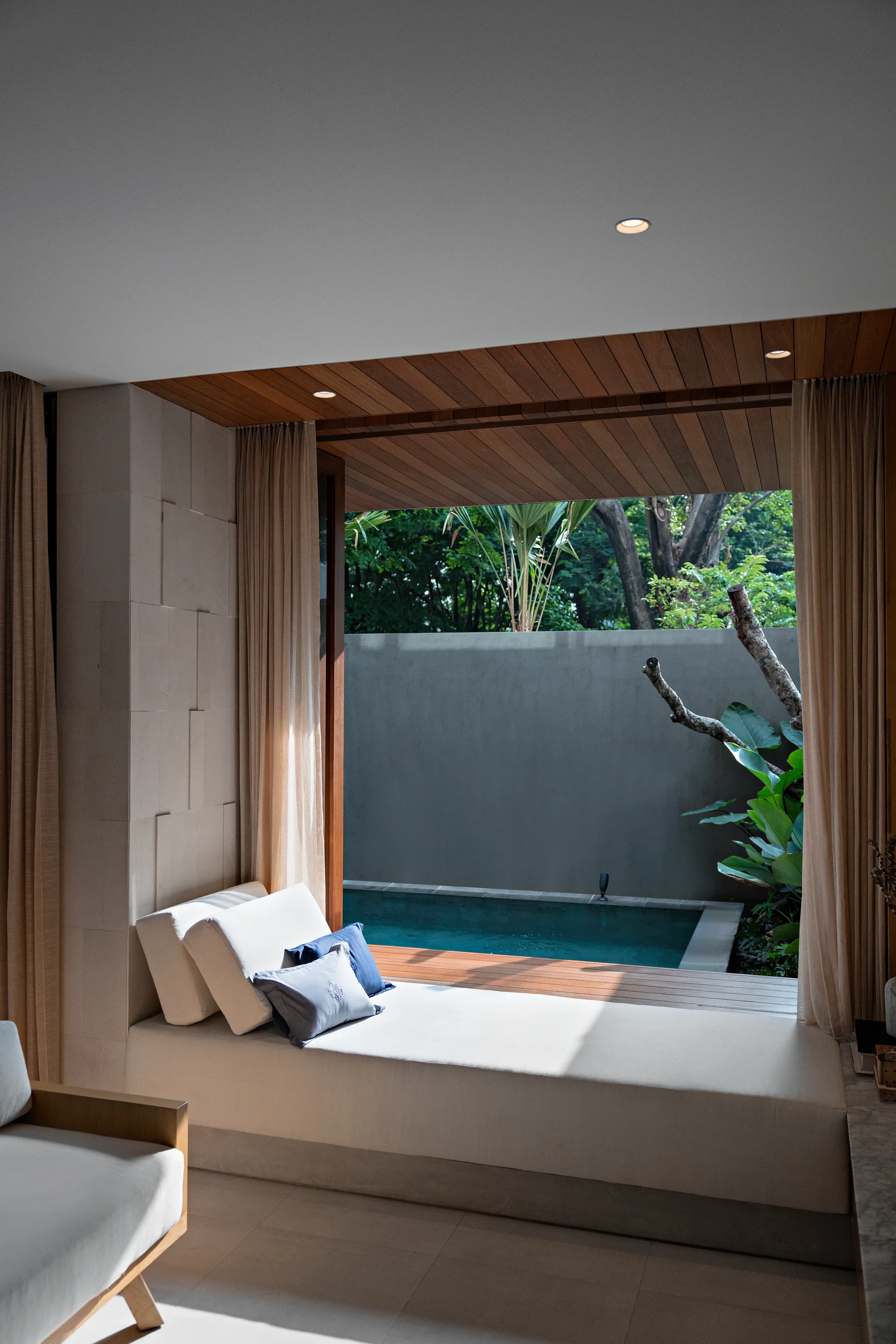 A minimalist indoor bedroom features a low daybed with white cushions next to open sliding doors that lead directly to a private swimming pool.
