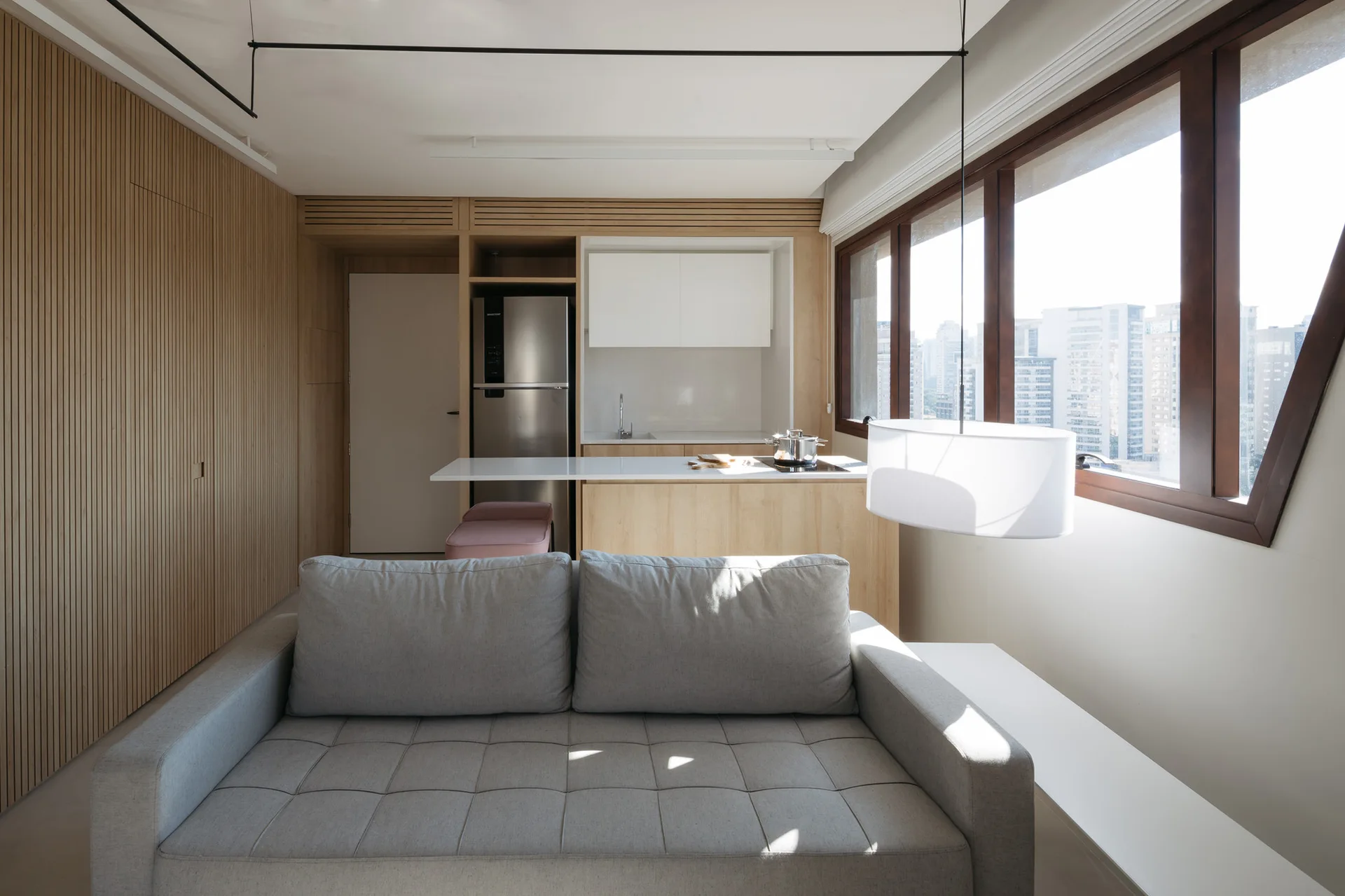 A compact open-concept living room and kitchen featuring a gray tufted sofa, a slatted wood accent wall, and large windows with a city view.