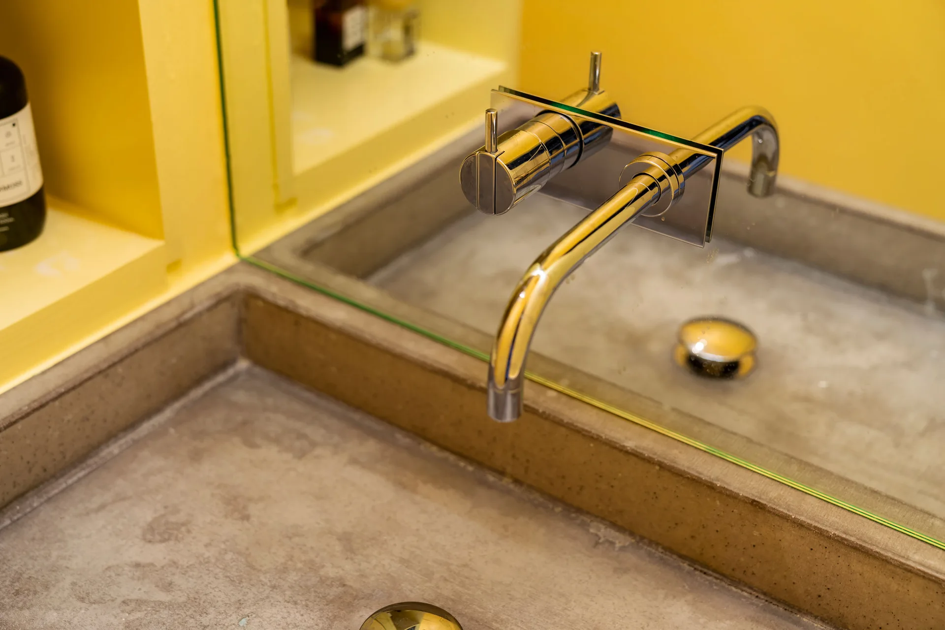 A wall-mounted polished chrome faucet is positioned over a textured grey concrete sink basin against a bright yellow wall and mirror.