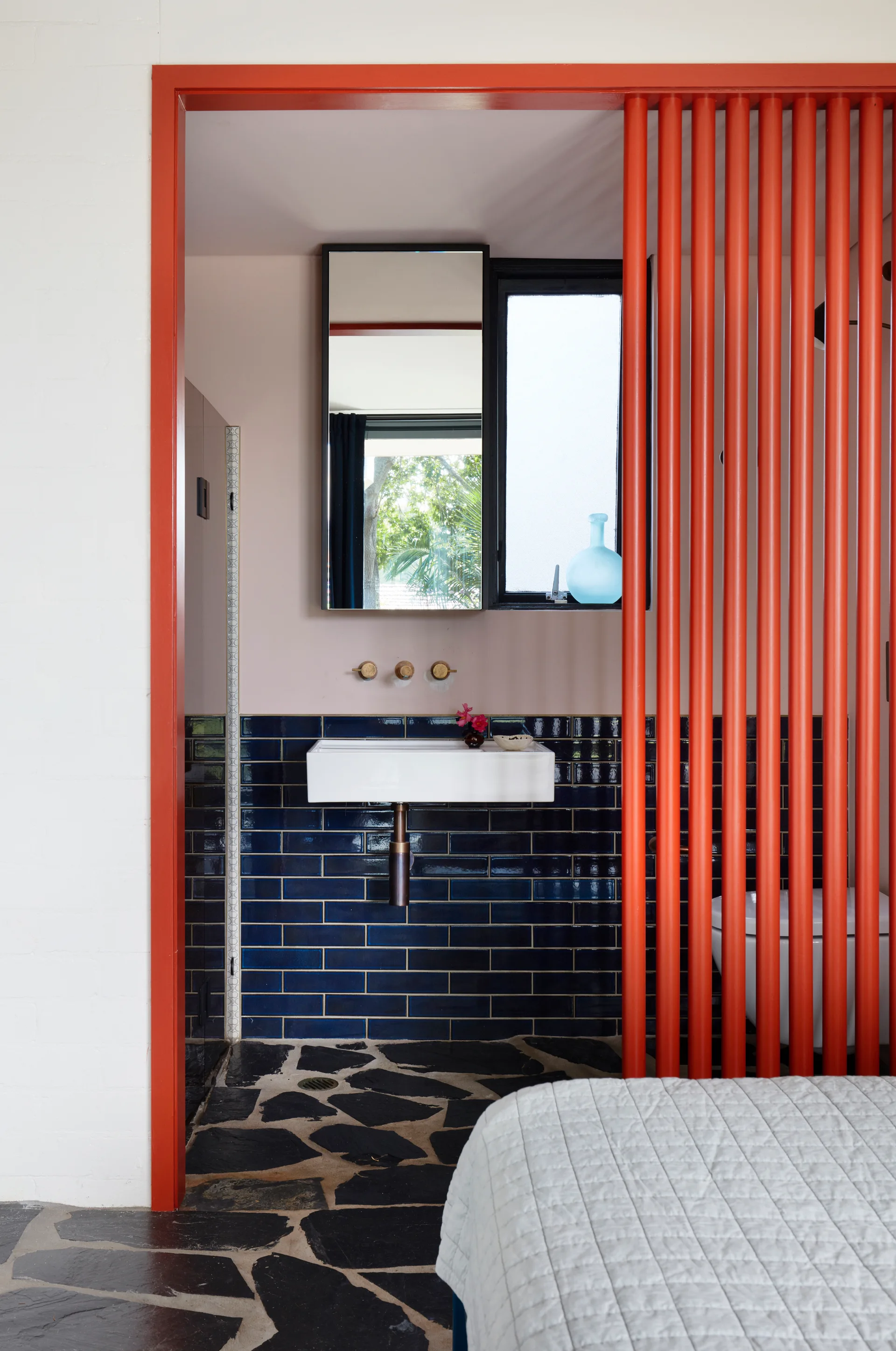 A modern bathroom viewed through a red frame, featuring a red vertical slat partition, blue subway tile wall, and flagstone floor.