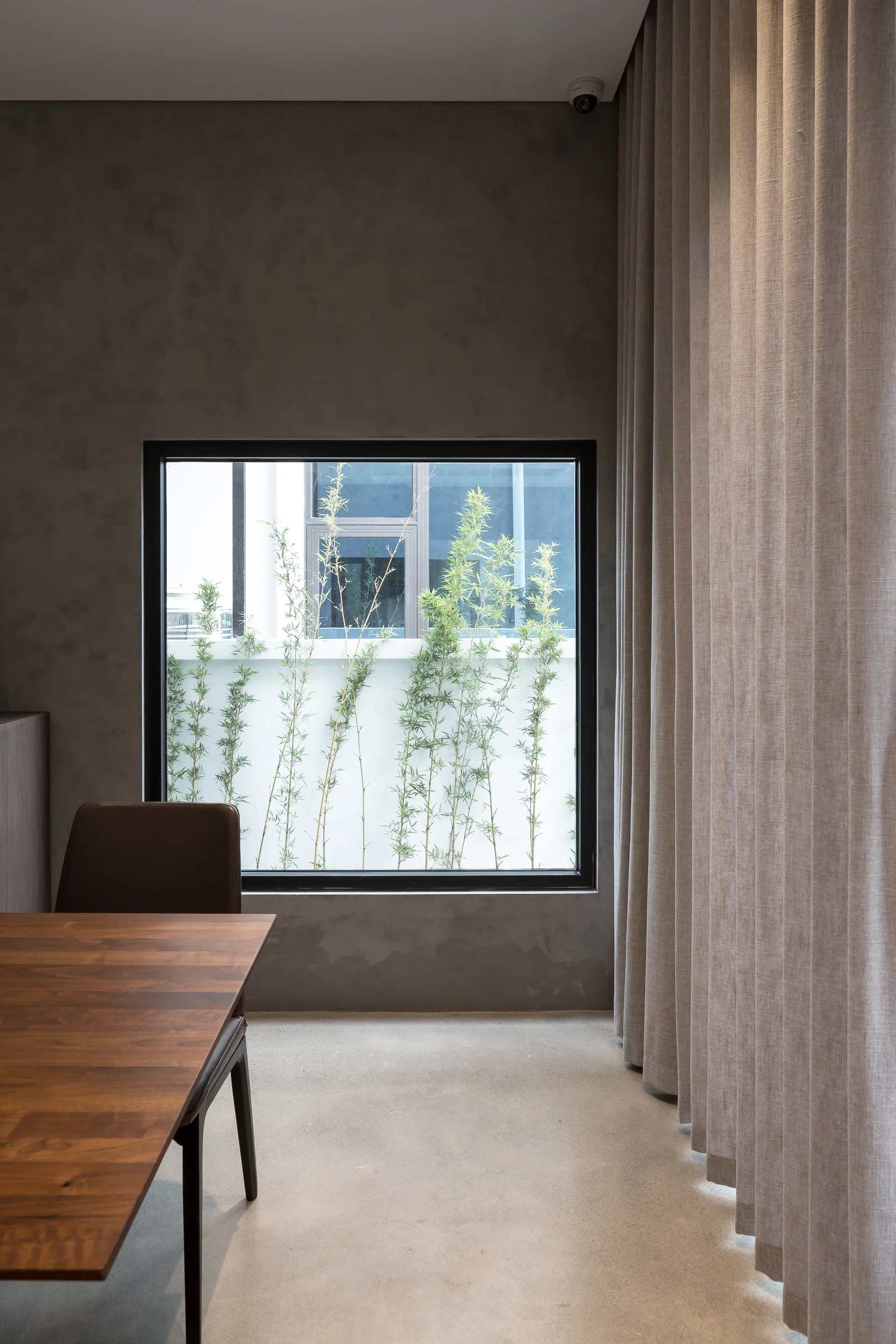 A minimalist dining area with a wood table, concrete walls, a large square window framing green bamboo, and floor-to-ceiling beige curtains.
