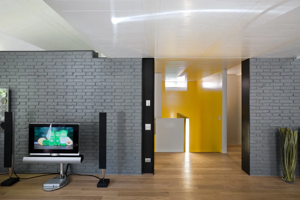 A living room features a grey painted brick wall, hardwood floors, a TV on a metal stand, and a bright yellow entryway in the background.