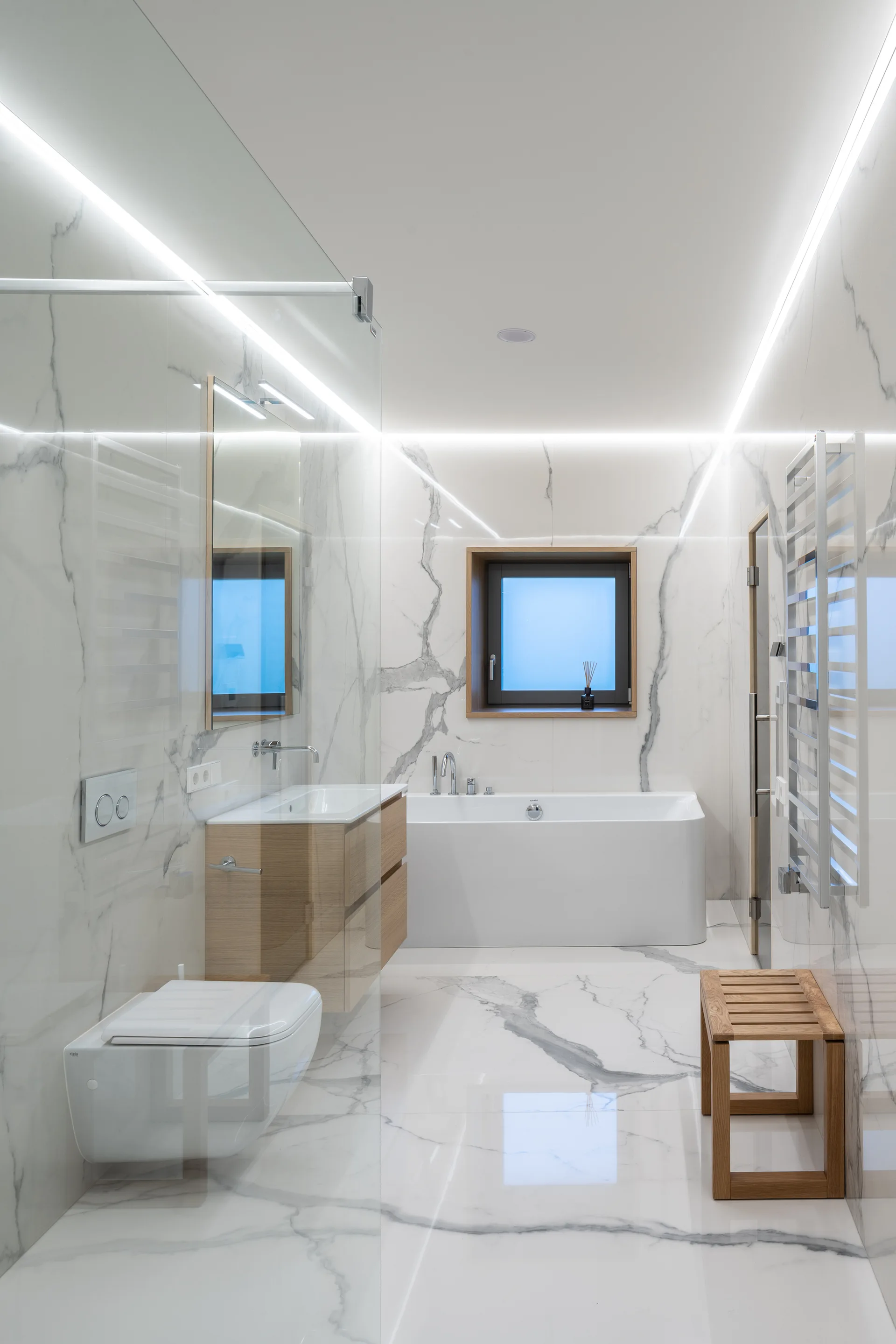 A modern bathroom lined in veined white marble tile features a floating wooden vanity, a wall-hung toilet, a white bathtub, and linear ceiling lights.