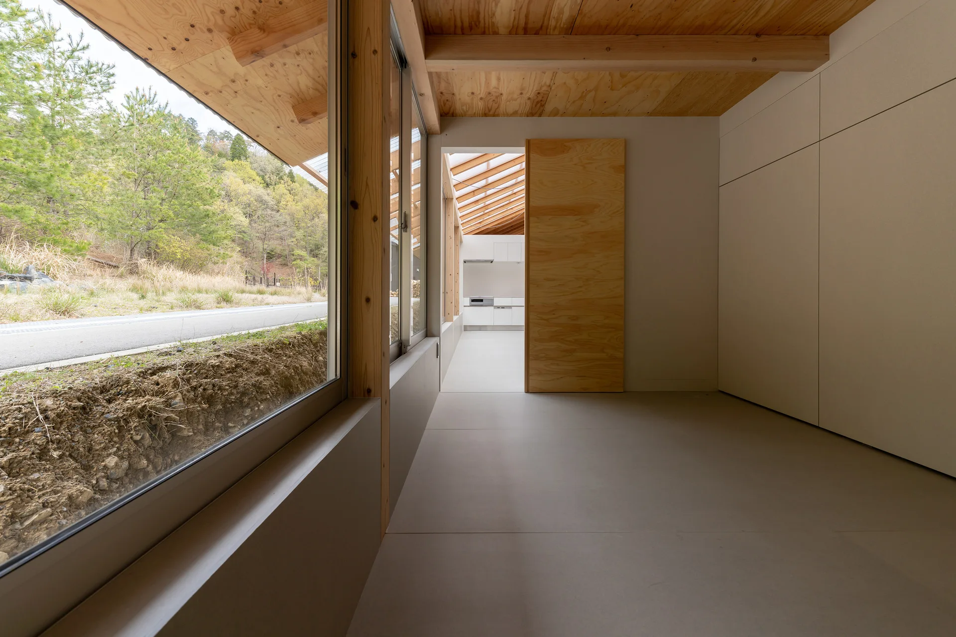 A minimalist hallway features a light plywood ceiling, a sliding wooden door, seamless wall cabinets, and a large window facing an outdoor landscape.