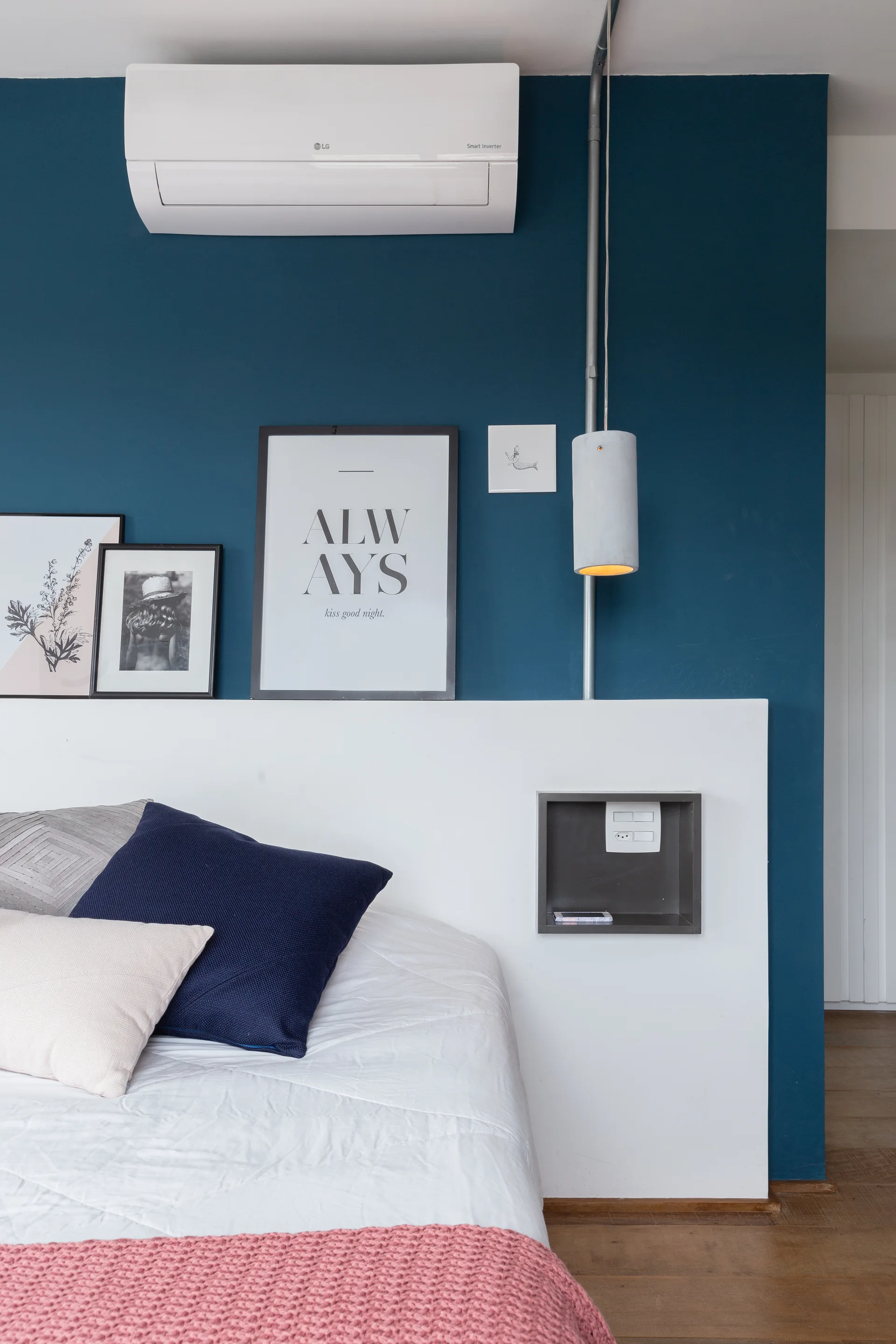 A white headboard panel set against a deep blue bedroom wall with framed art, a pendant light, and a built-in shelf.