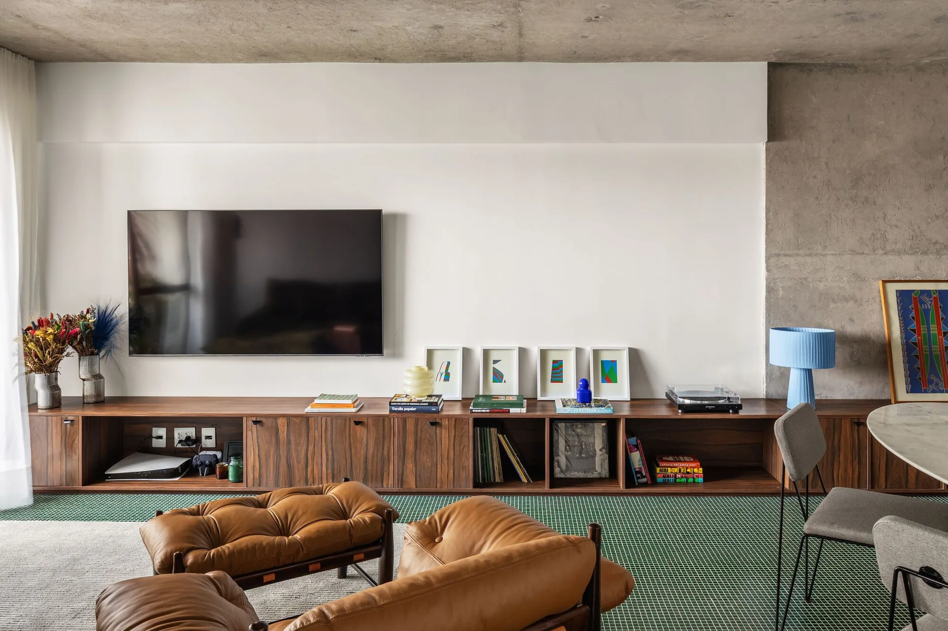A modern living room features a low wood media console beneath a wall-mounted TV, tufted leather armchairs, and a green grid-patterned tile floor.