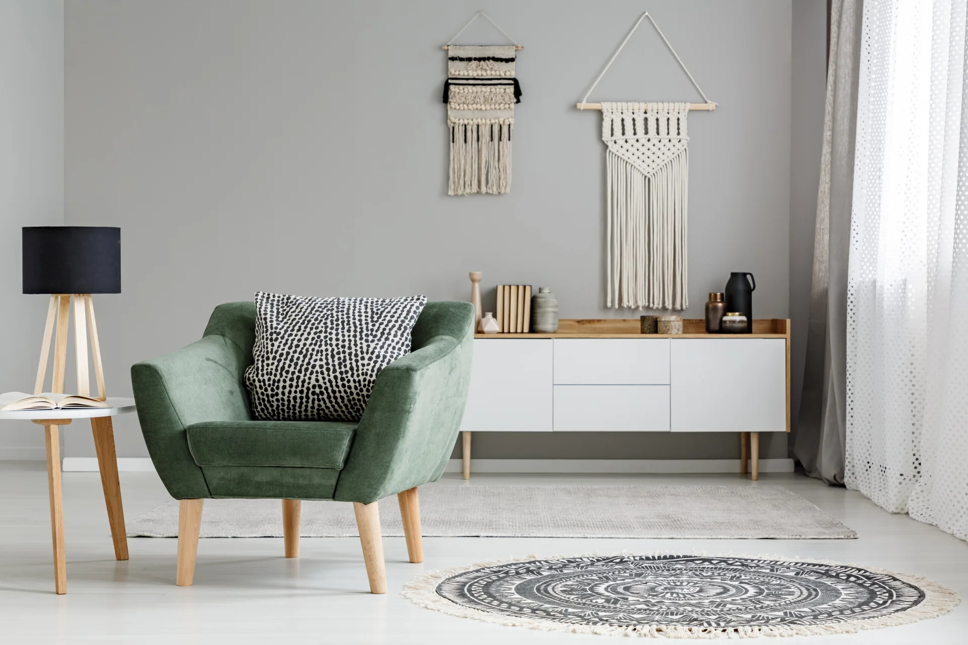 A sage green armchair with a patterned pillow sits next to a white credenza with macrame wall hangings on a gray wall.